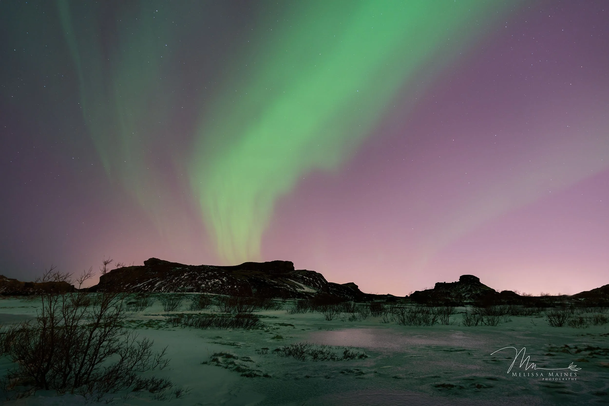 Nighttime Icelandic landscape showing green and pink northern lights with snow-covered ground underneath.