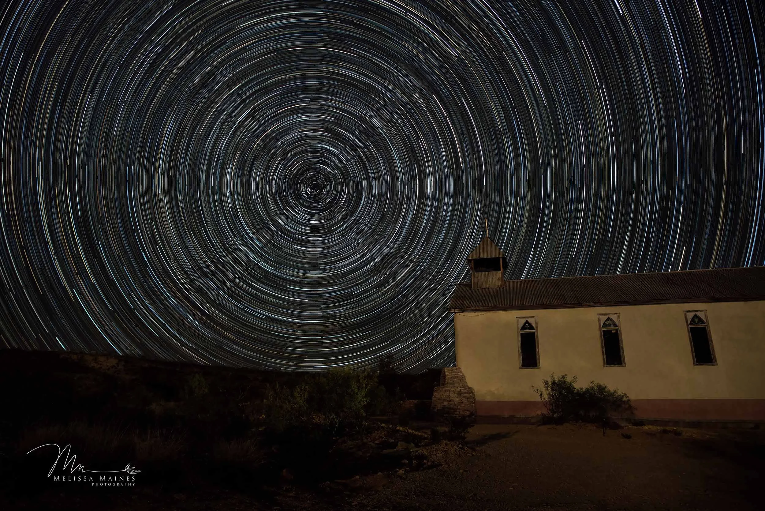 Long exposure photograph of star trails over an old church with a rusted roof and arched windows in a dark landscape.