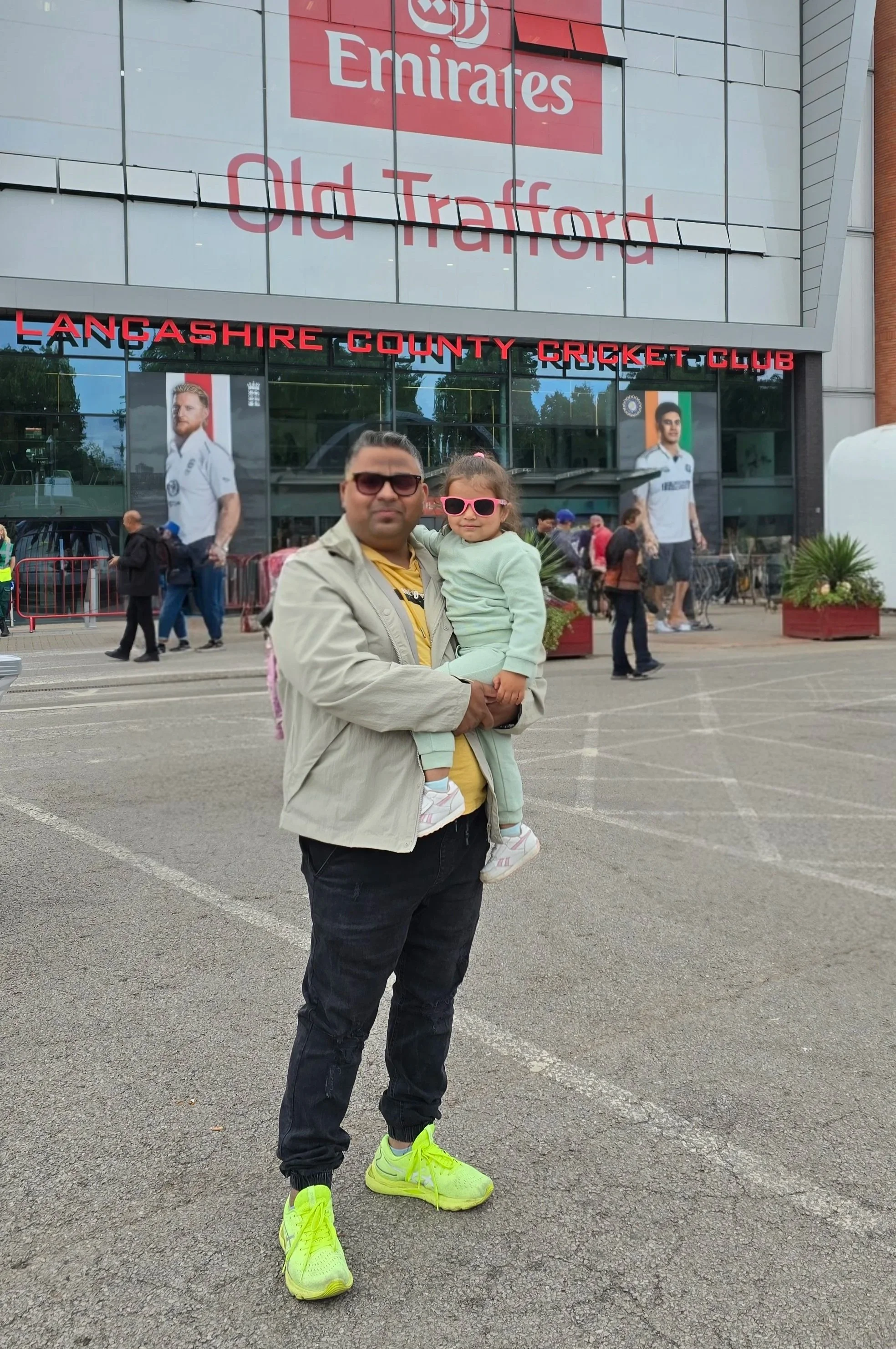 A man is holding a young girl in front of Langashire County Cricket Club and Old Trafford stadium with people walking by.