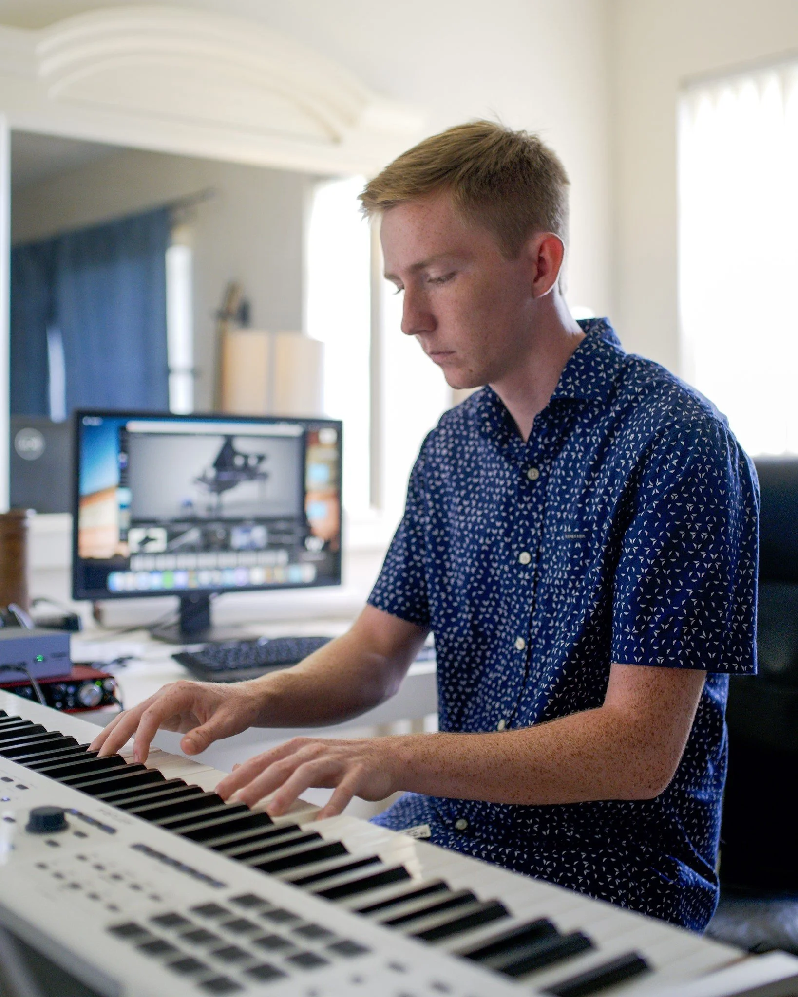 Young man playing keyboard in a home studio with computer and monitor in the background.