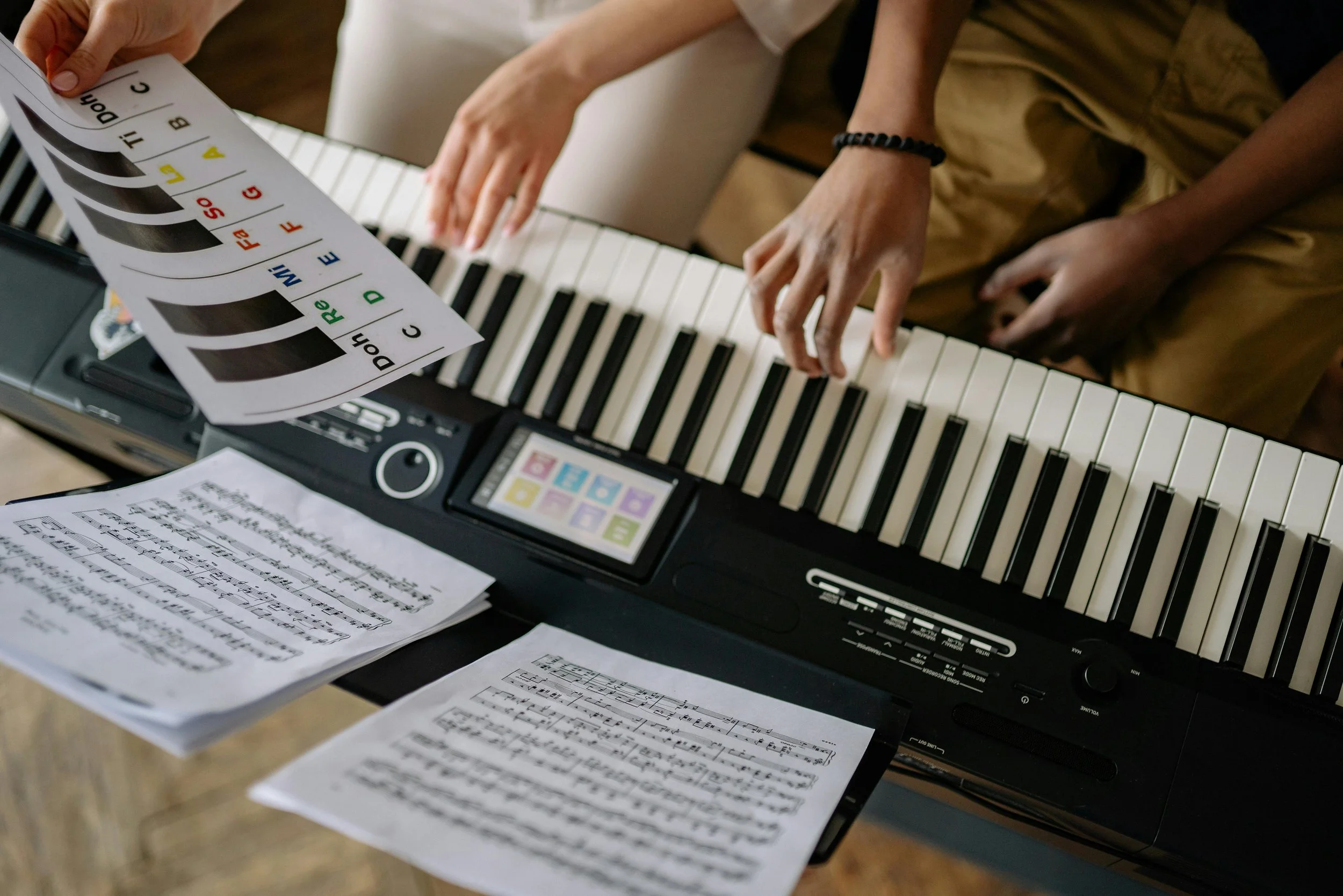 A person sitting at a digital piano, with sheet music and a colored music theory chart or guide. The person’s hands are on the keys, and the scene includes musical notes and symbols.