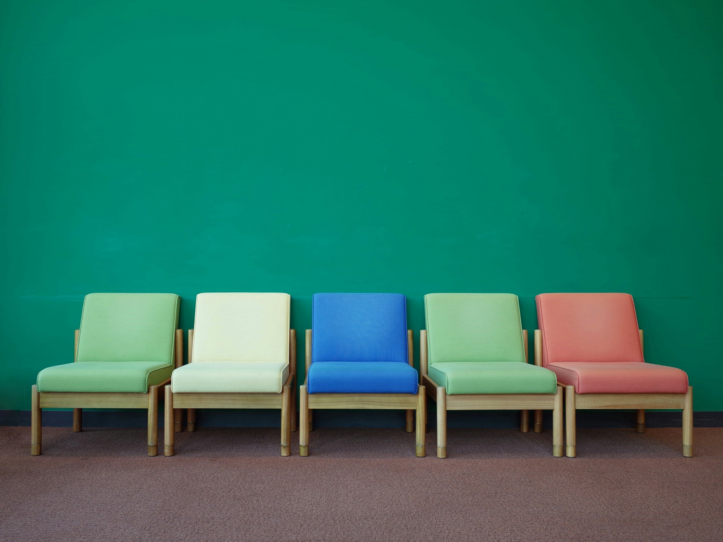 Six colorful chairs arranged in a row against a green wall, with a brown carpeted floor.