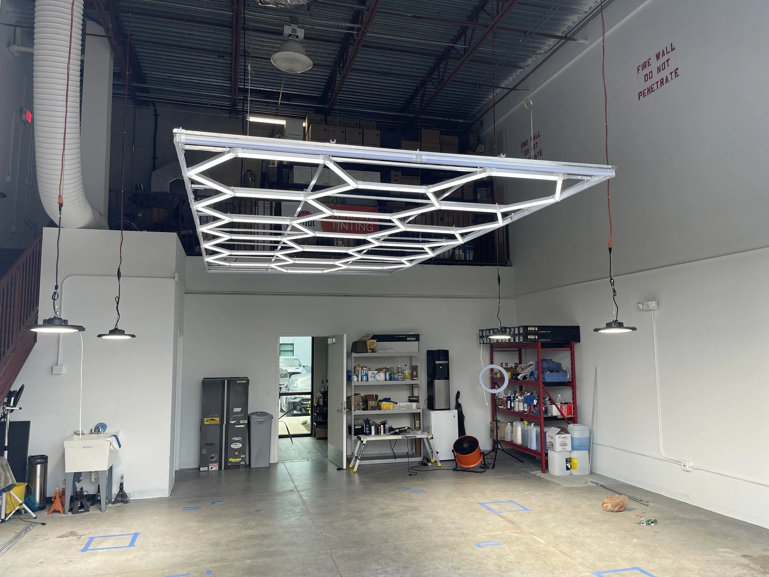 Industrial space with a modern geometric ceiling light fixture, white walls, and a mix of storage shelves, tools, and equipment near an open door leading outside.