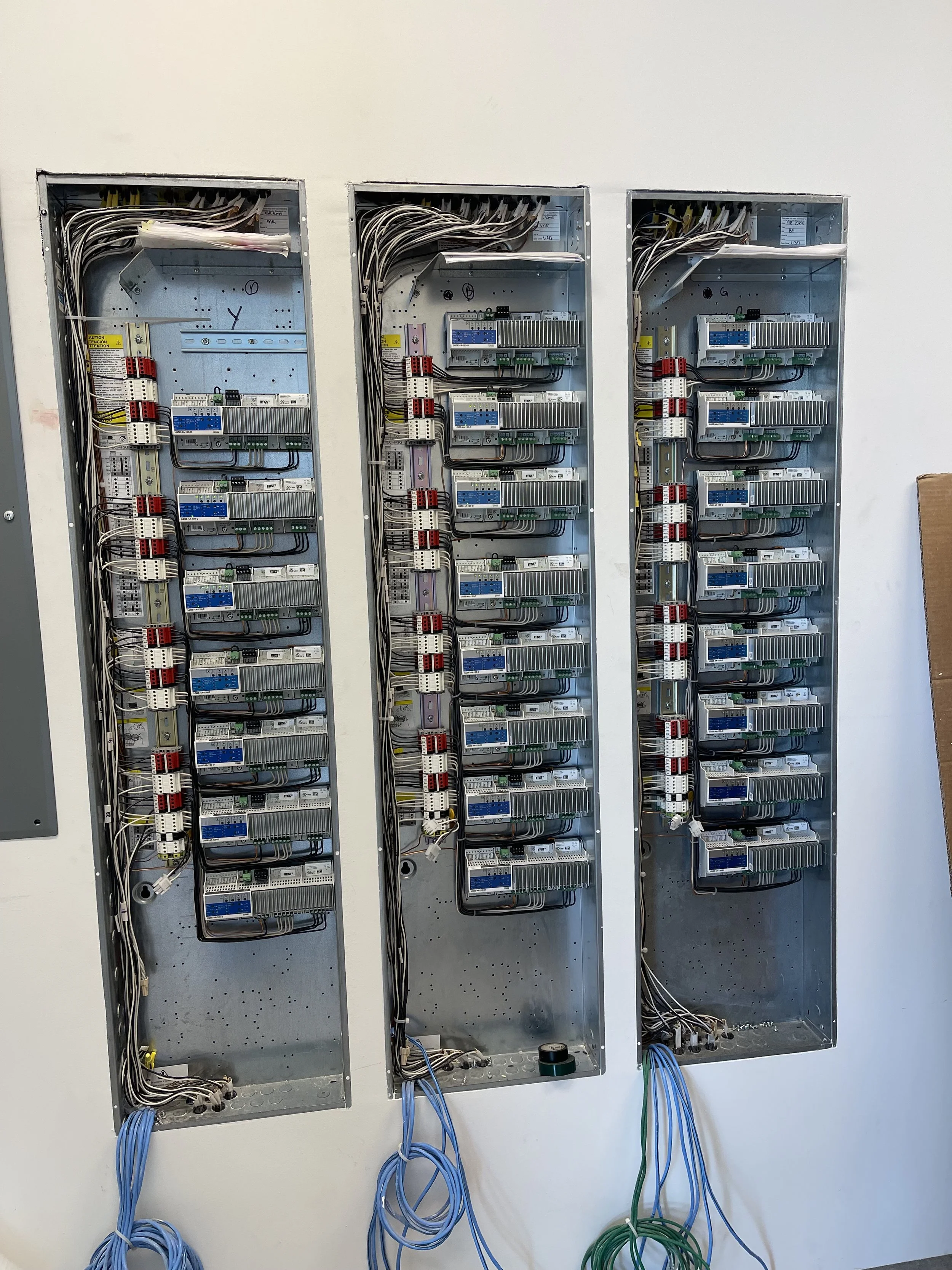 The image shows three opened electrical panels with wiring and components inside, mounted on a white wall.