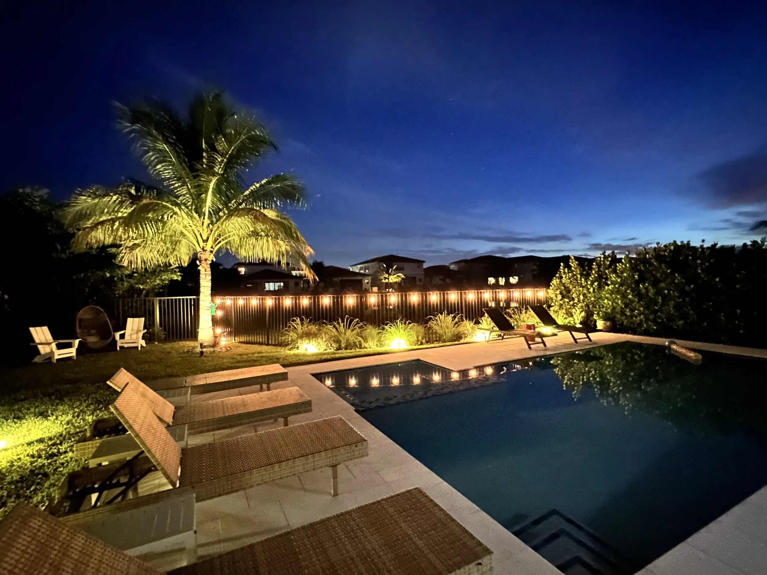 Nighttime view of a backyard pool area with lounge chairs, a tall palm tree, and outdoor lighting, overlooking rooftops and a darkening sky.