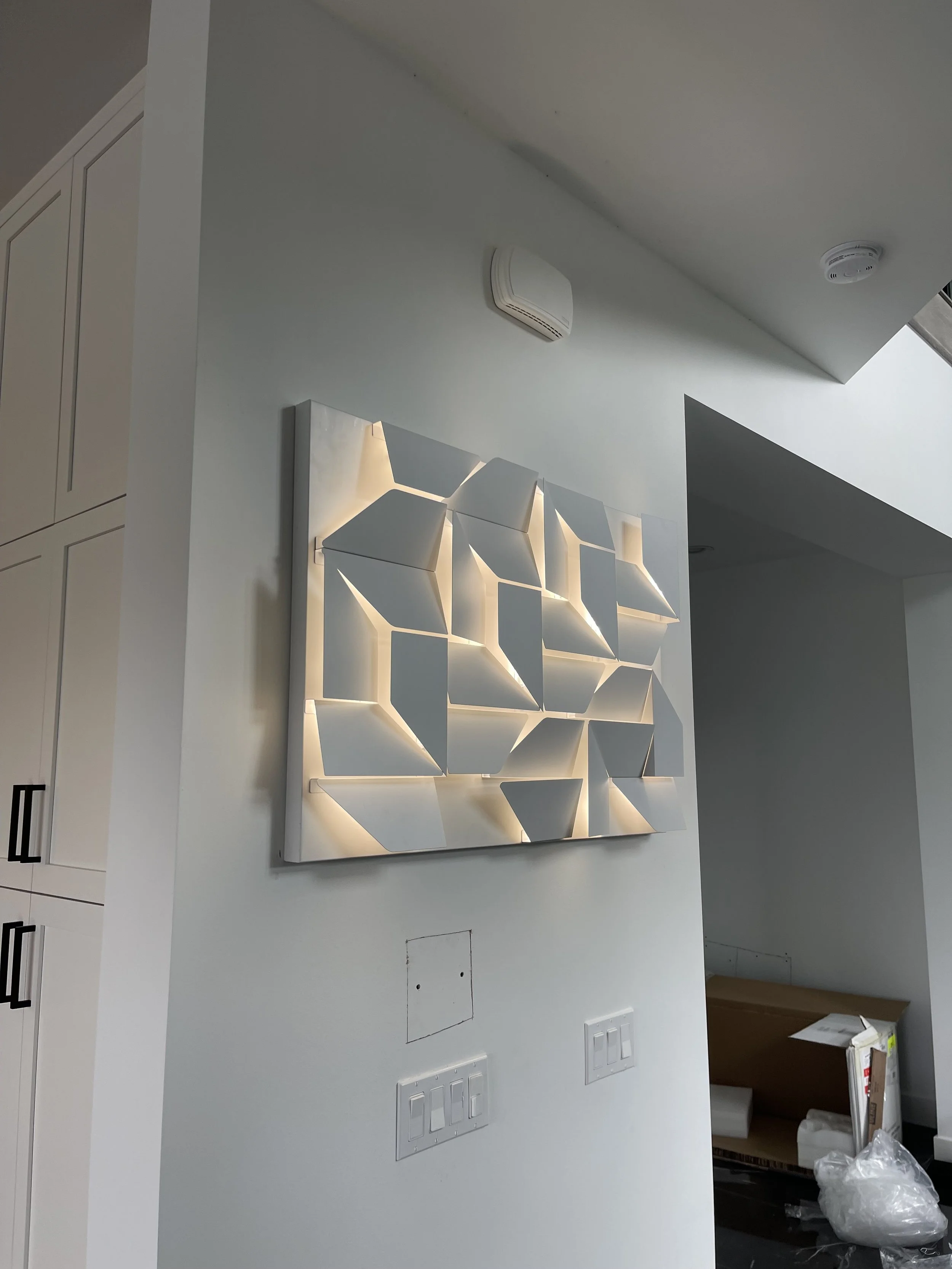 Modern wall art with geometric white panels illuminated by backlighting, mounted on a white wall.