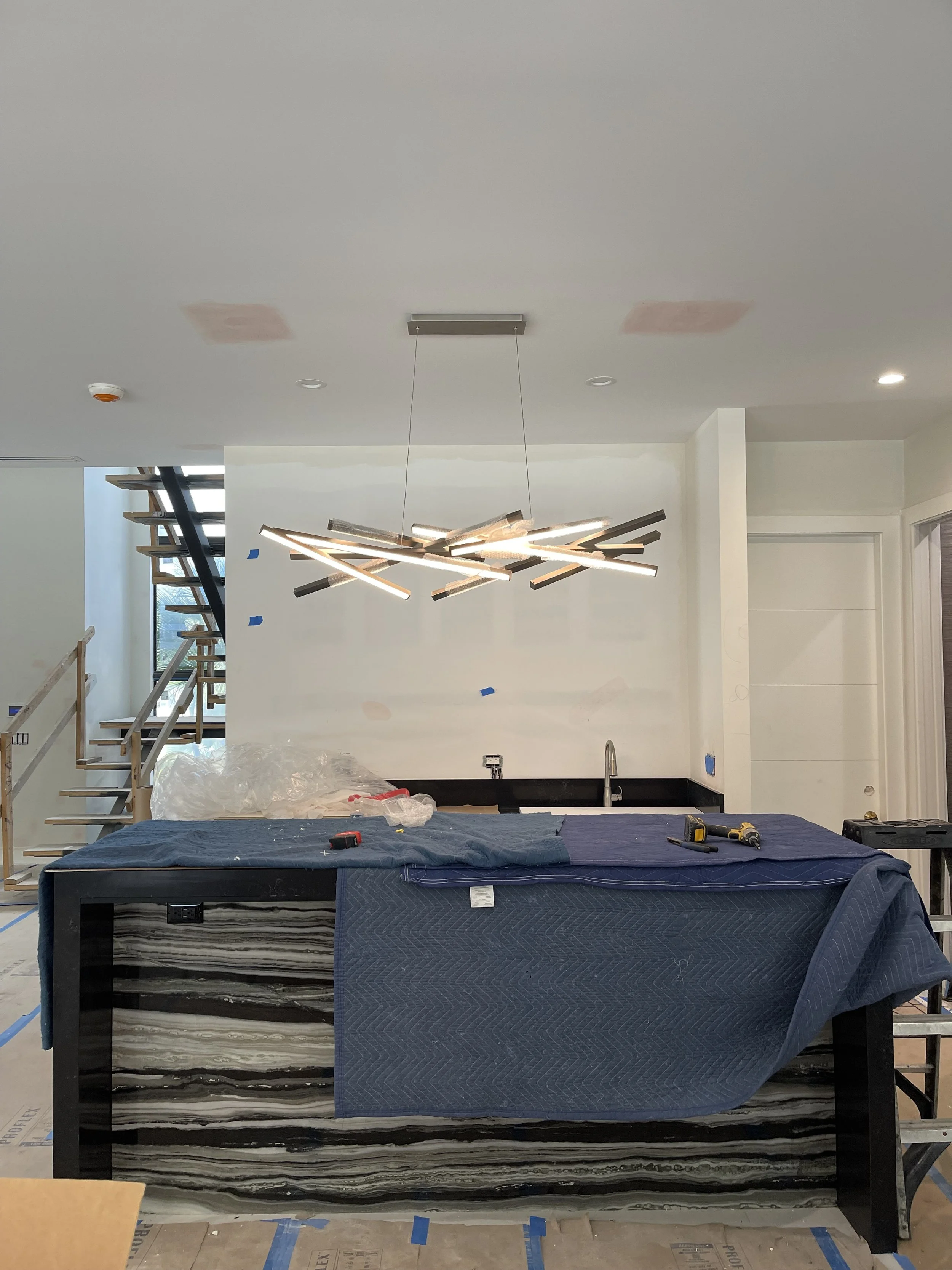 Modern kitchen under construction with a black marble island, construction tools on the surface, an artistic chandelier hanging from the ceiling, and a staircase leading to the upper floor. The walls are partially painted.