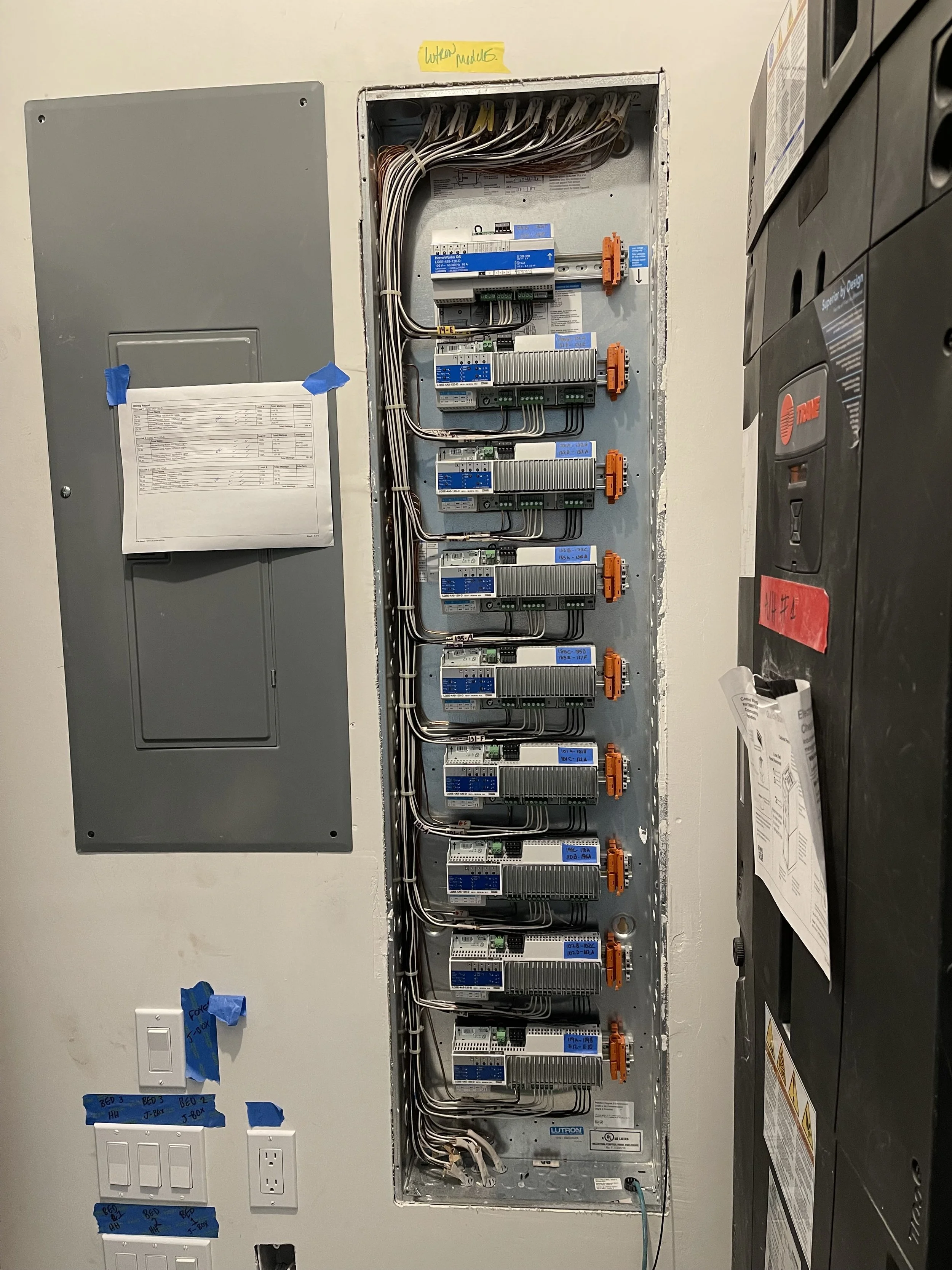 Electrical panel with multiple circuit breakers and wires, mounted on a wall in an electrical room.