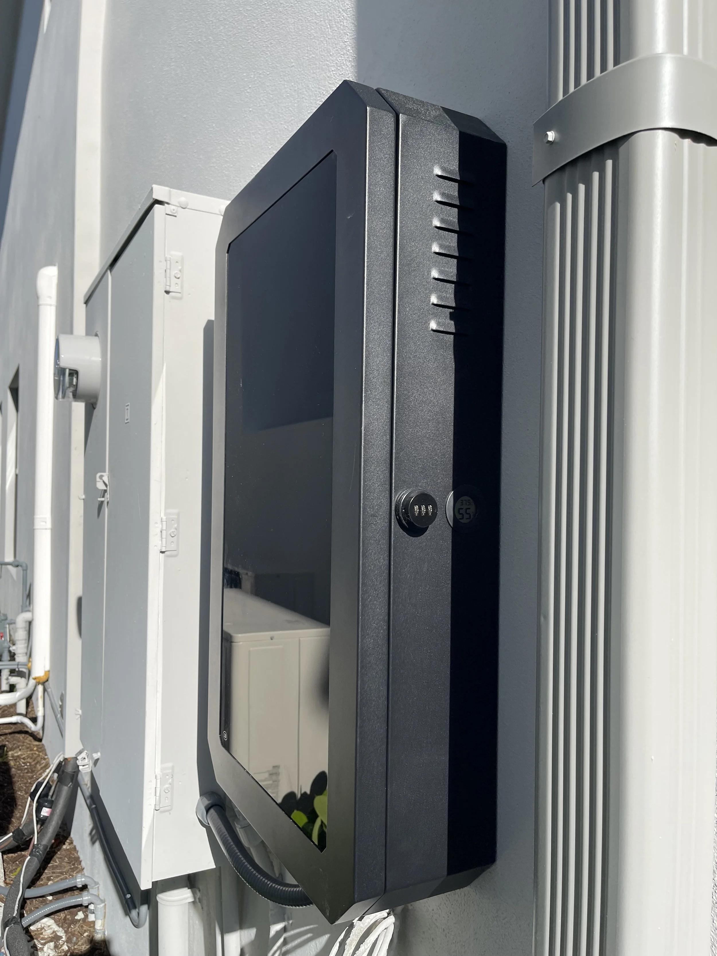 A black electronic device, possibly a security monitor or control panel, mounted on an exterior wall, with two digital displays showing the temperature and other information.