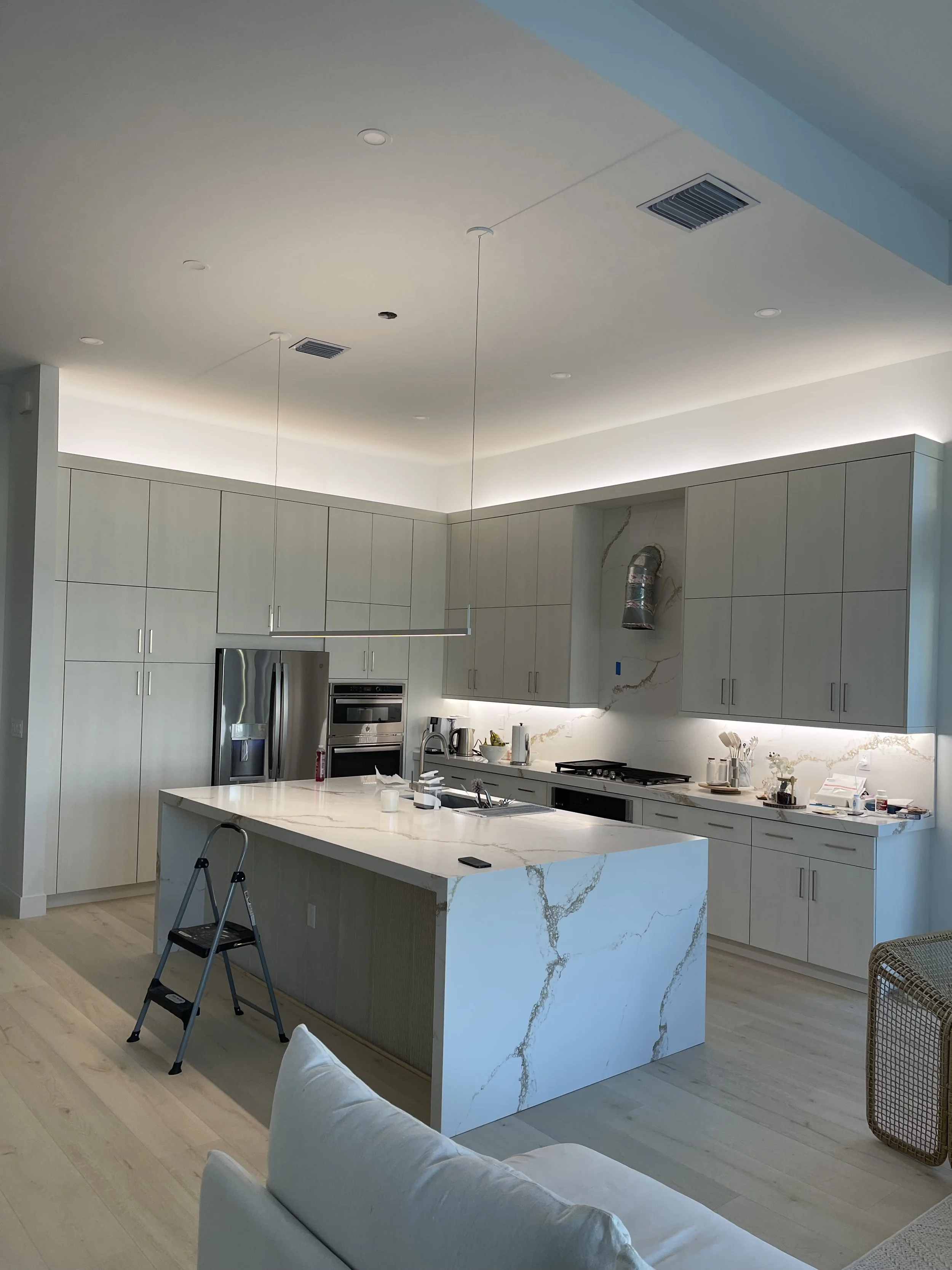 Modern kitchen with light gray cabinets, a large marble island, stainless steel refrigerator, built-in oven, and minimal clutter, with a step stool near the island.