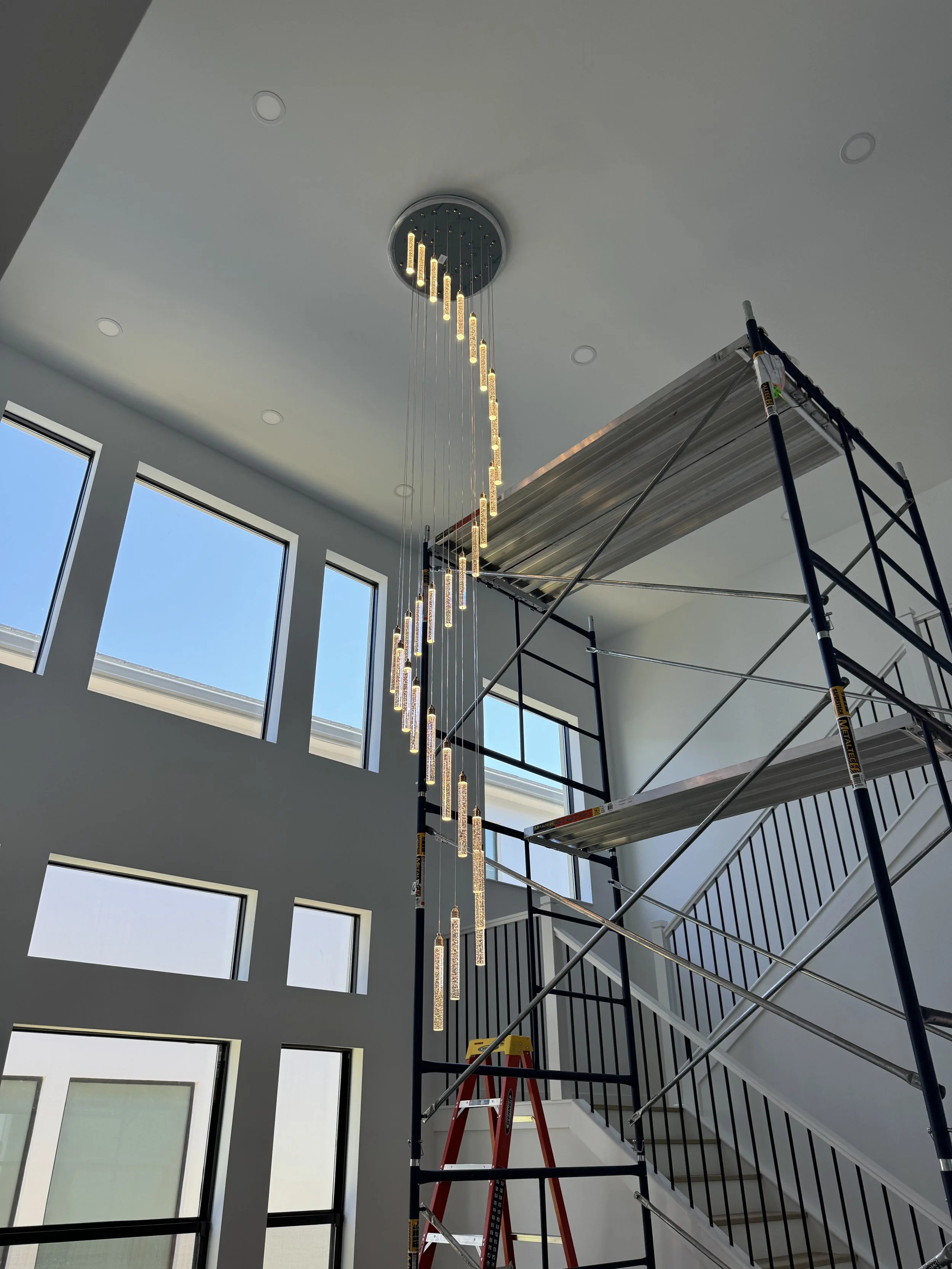Interior view of a modern house with large windows, a hanging chandelier, a staircase with black railings, and a scaffolding platform.