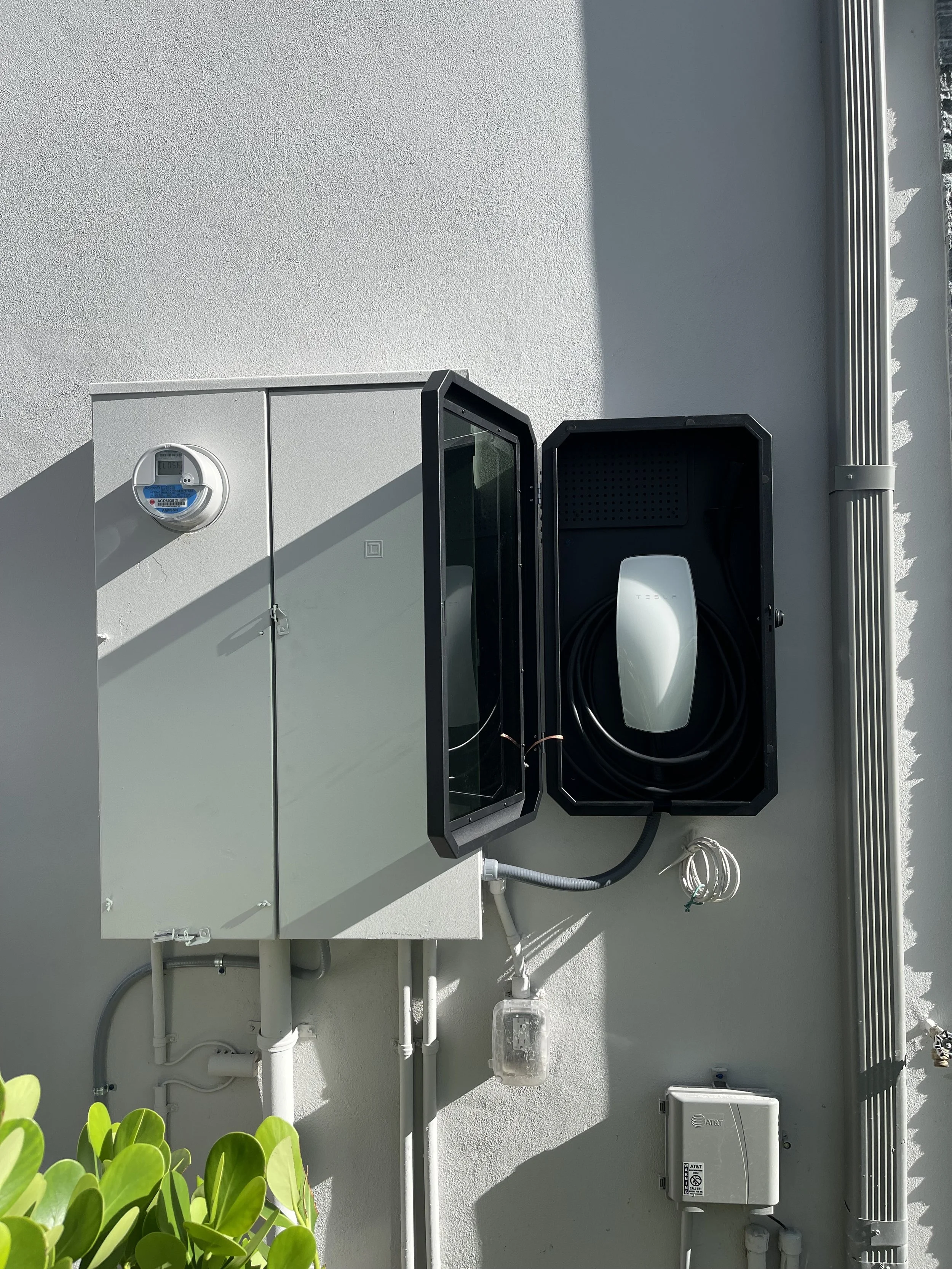 An electric vehicle charging station installed on an exterior wall with a Tesla charging unit inside an open black case, adjacent to a utility meter and various electrical boxes.
