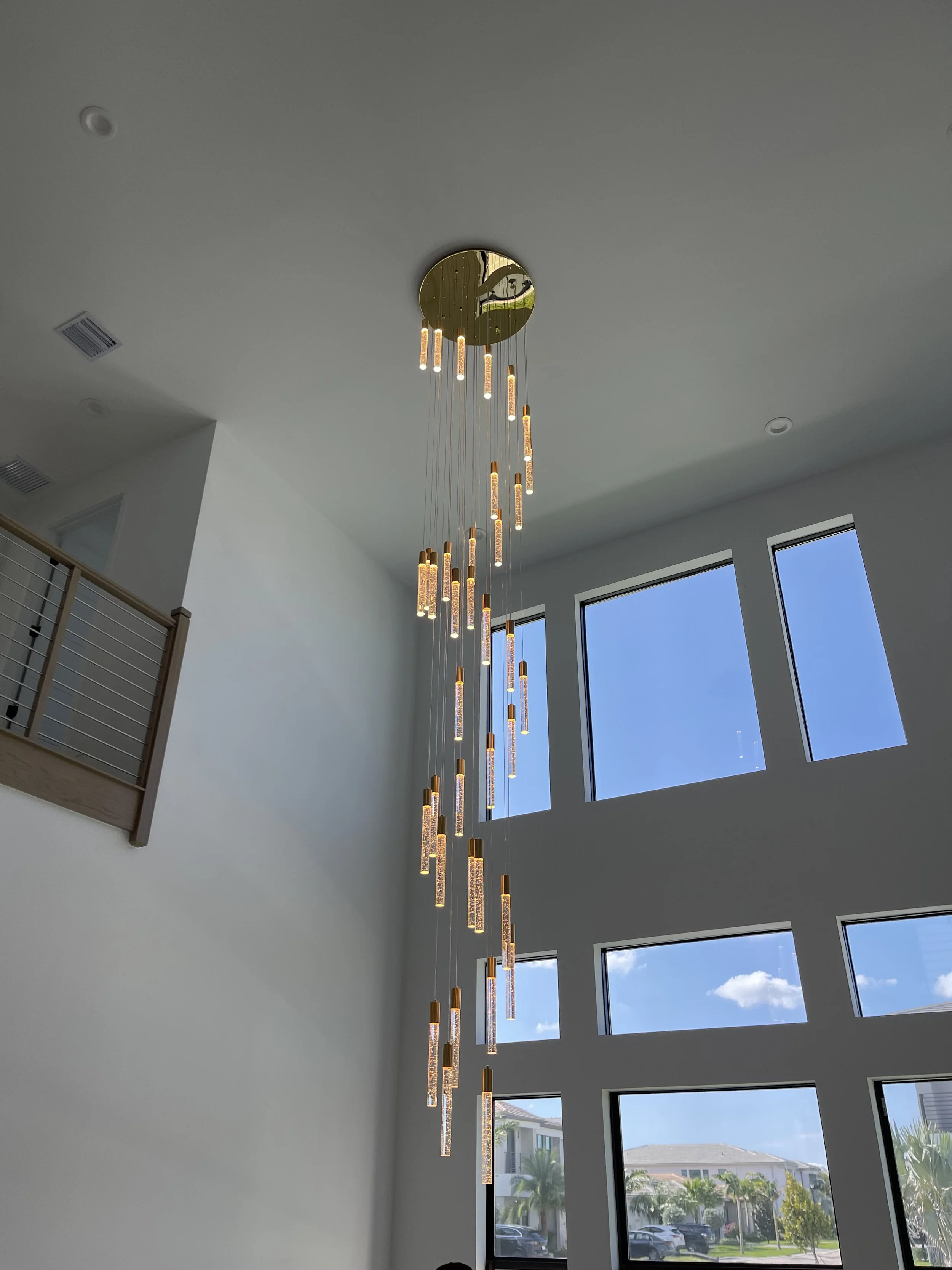 Modern chandelier with multiple hanging LED light tubes suspended from a reflective gold circular ceiling mount in a bright two-story room with large windows and a view of neighborhood houses.