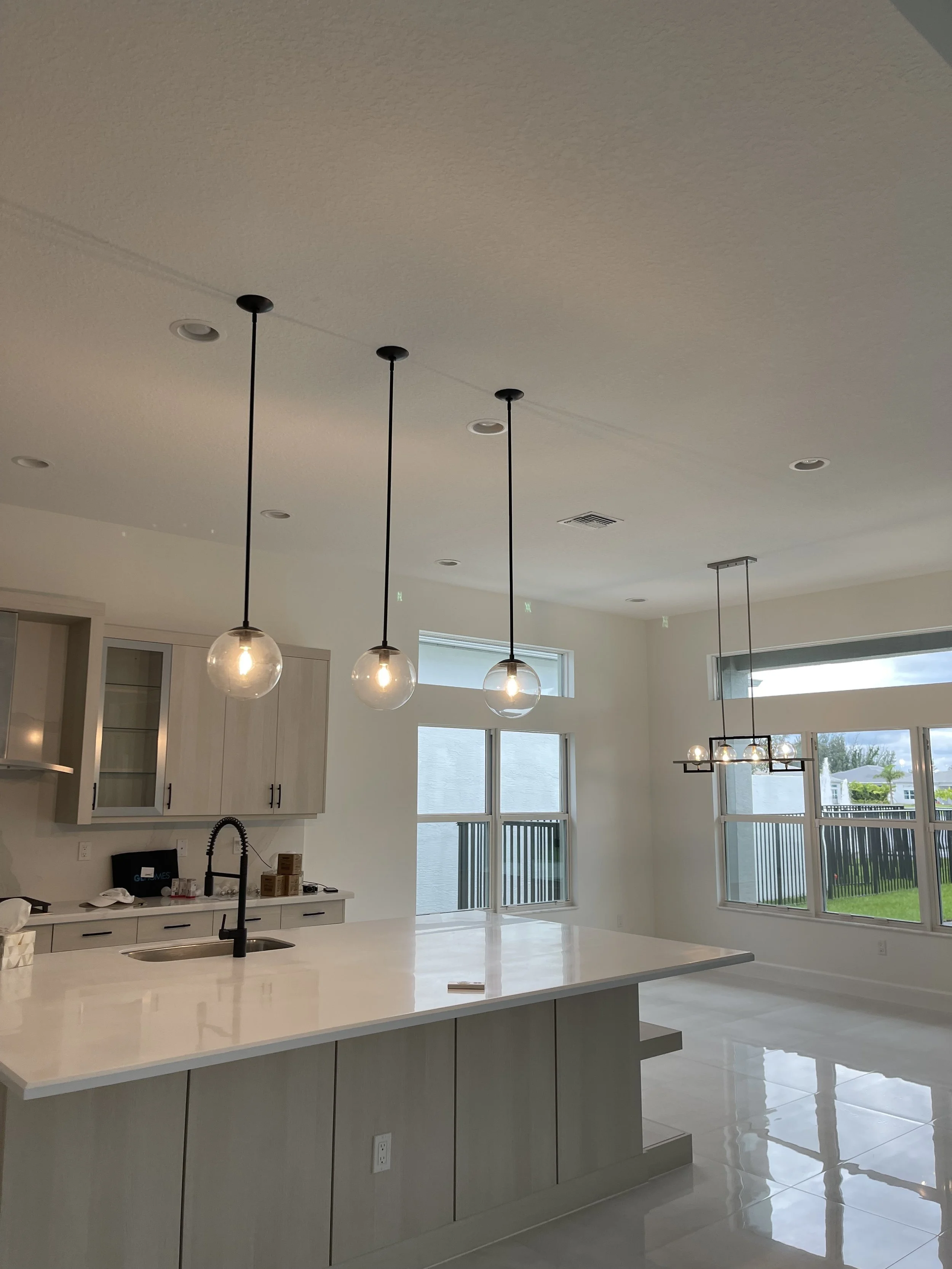 Modern kitchen with white cabinetry, a large island with a white countertop, black faucet, and three pendant lights above the island. Bright windows reveal a backyard with green grass and a black fence.