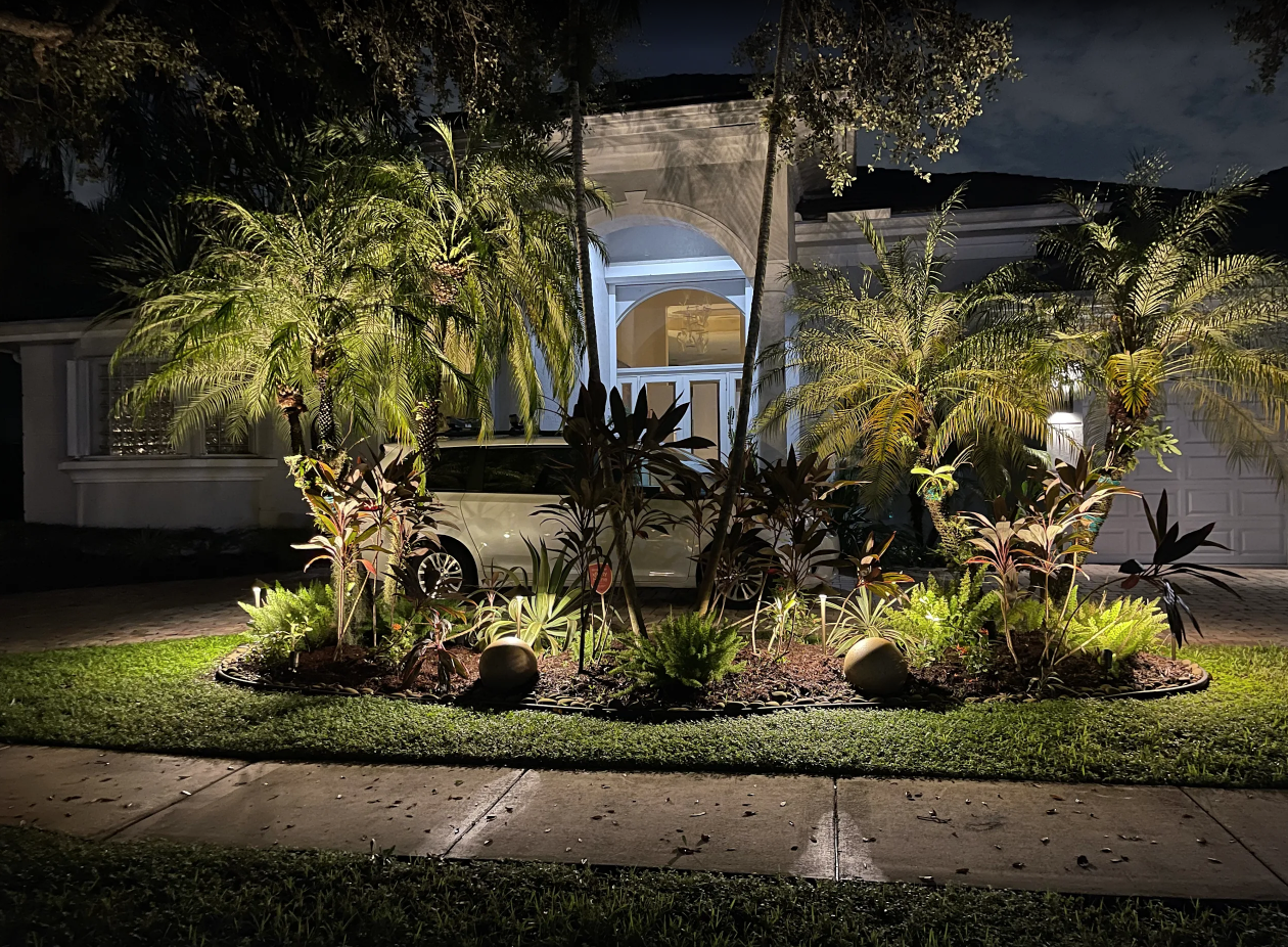 Night view of a landscaped front yard with tropical plants and palm trees illuminated by outdoor lighting, with a white vehicle parked near the house in the background.