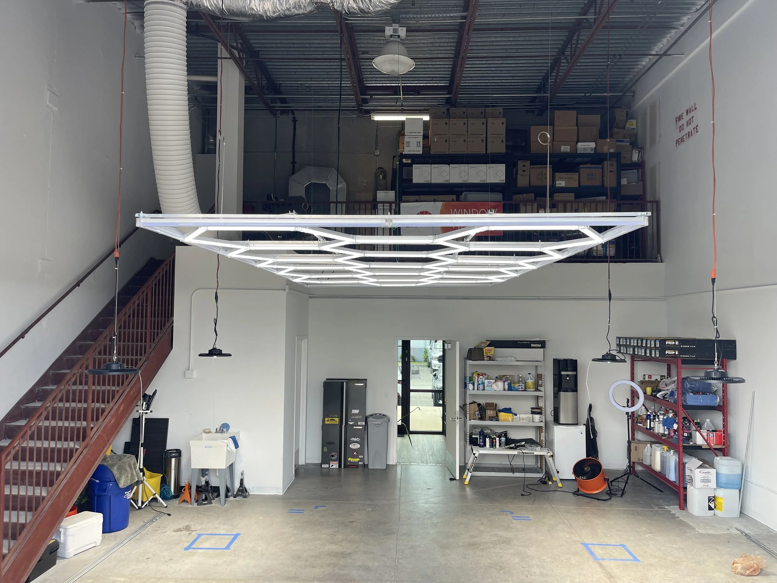 Interior of a commercial or industrial space with storage shelves, equipment, and tools. There are boxes on a mezzanine level, a large lighting fixture hanging from the ceiling, and various supplies on shelves and the floor.