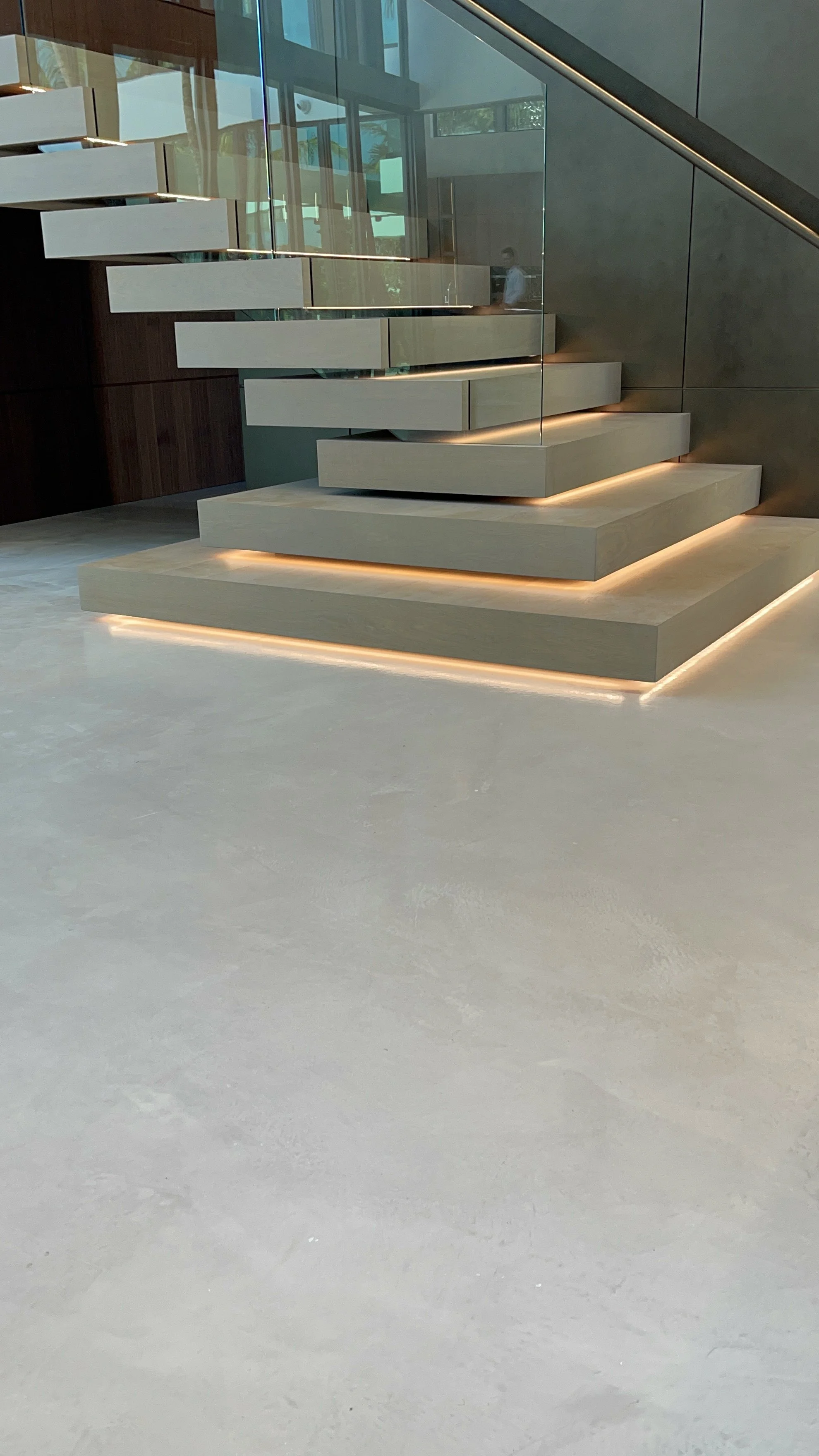 Modern interior staircase with beige steps, embedded LED lighting, glass railing, and large windows in a contemporary building.