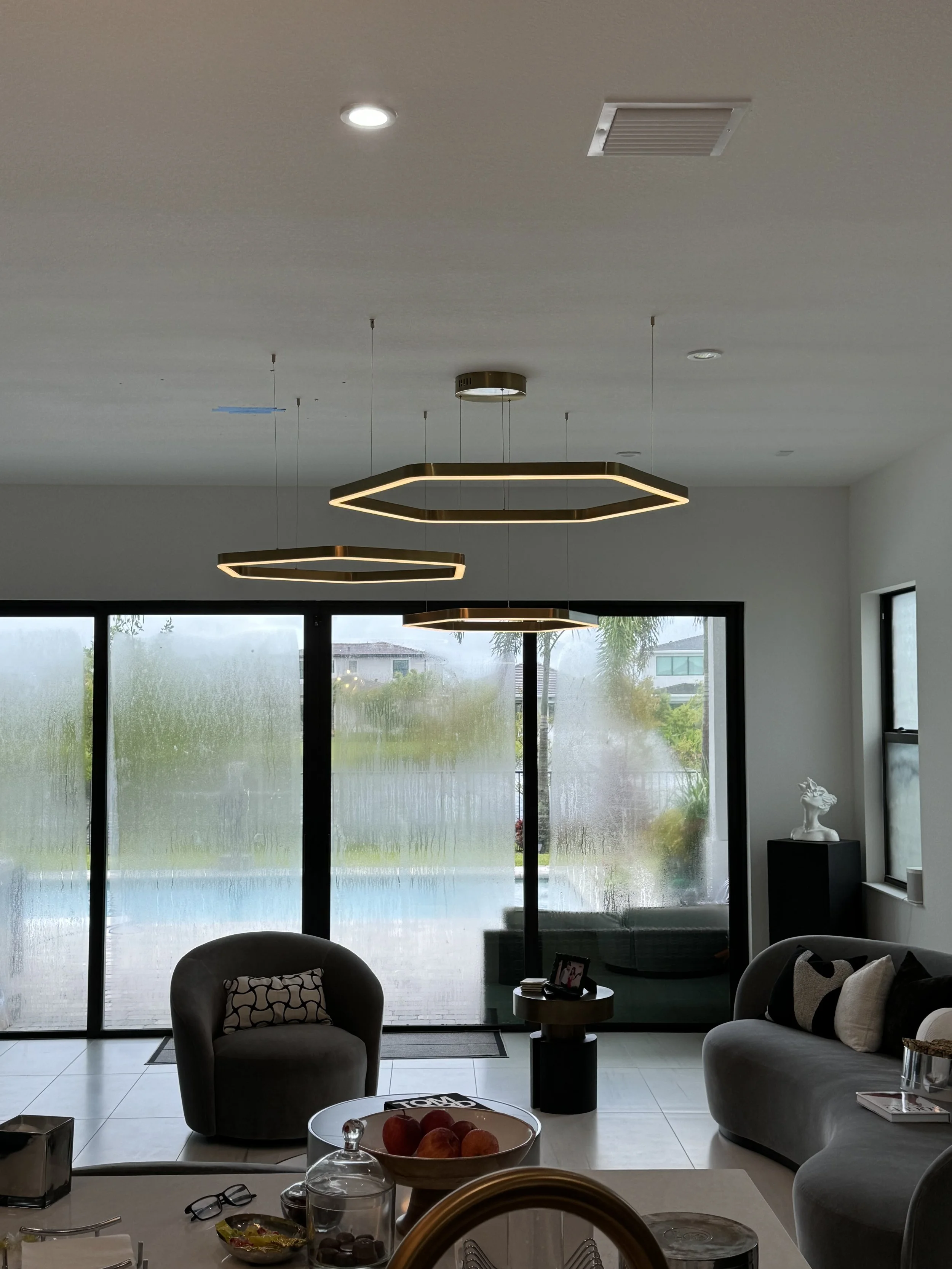 Modern living room with large glass sliding doors, contemporary ceiling lights, gray chairs, sofa with pillows, small side table, and decorative items.