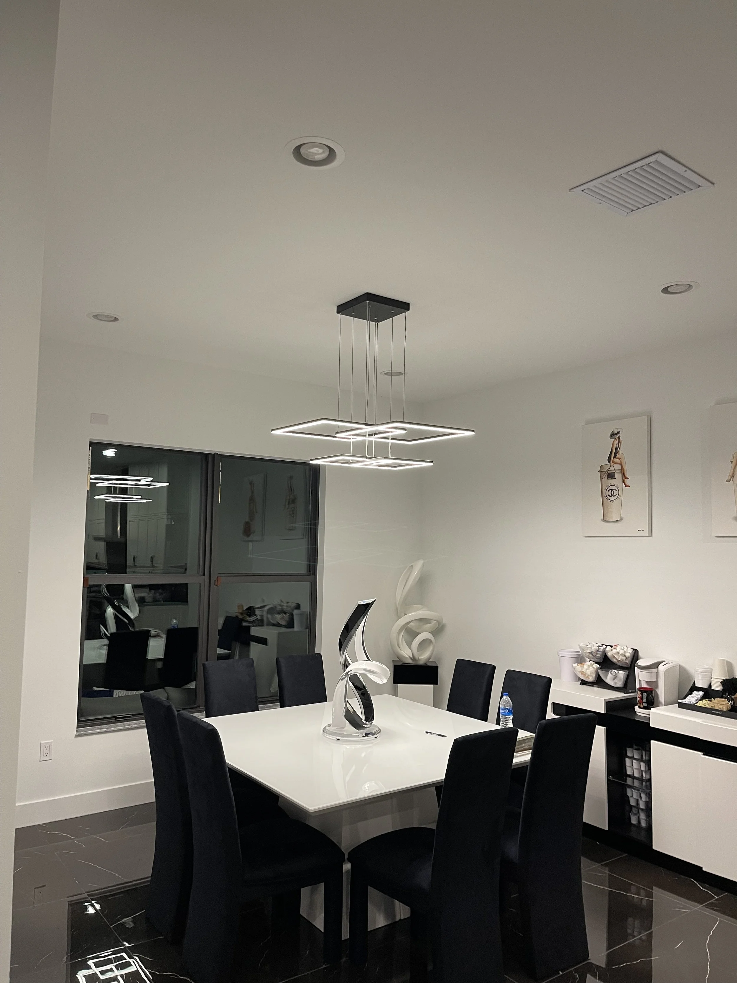 Modern dining room with a white rectangular table, black chairs, white walls, large windows, abstract sculptures, and artwork.