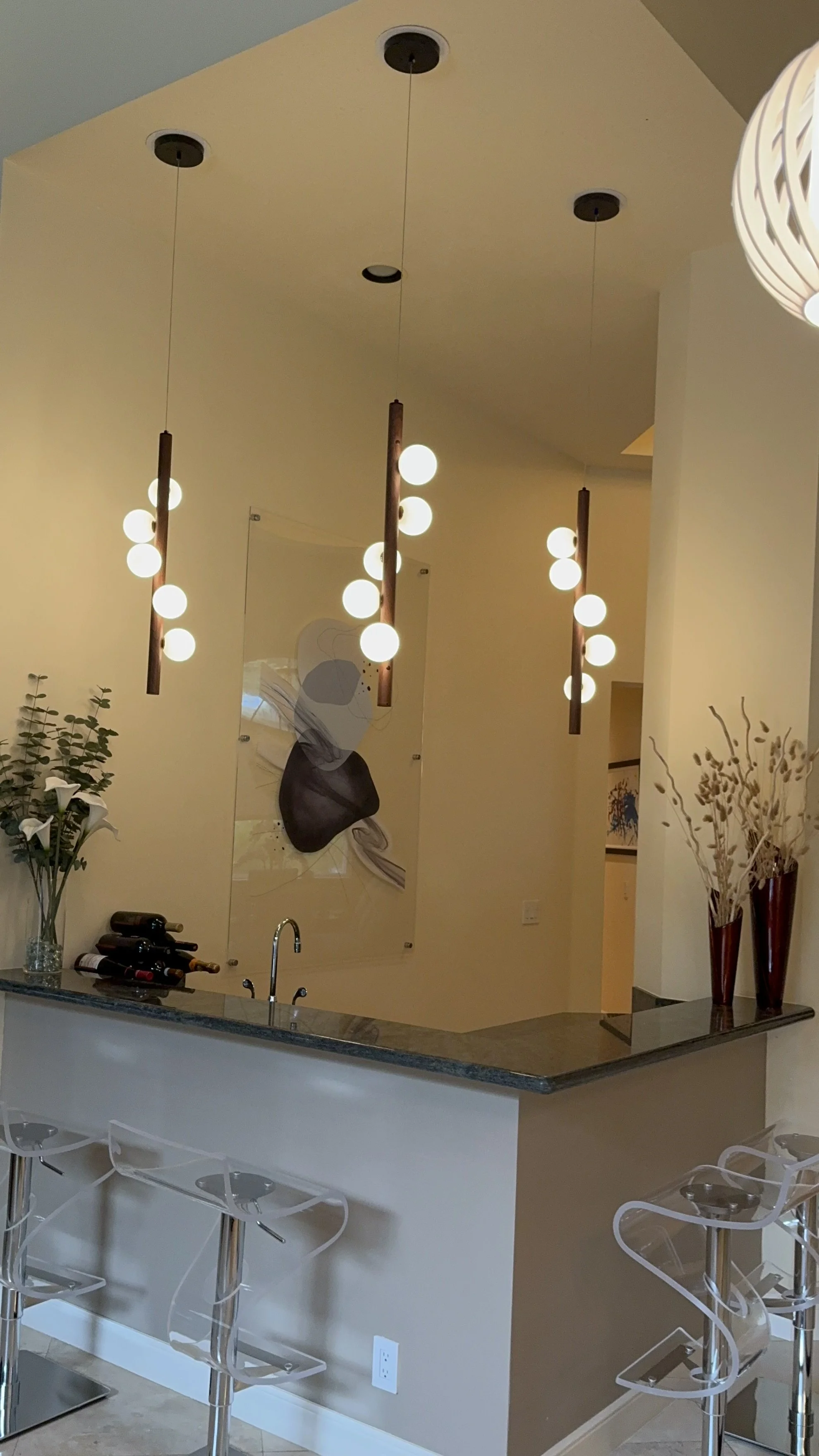 Modern kitchen with hanging pendant lights, a granite countertop with a sink, wine bottles on the counter, and decorative vases with plants and dried flowers.