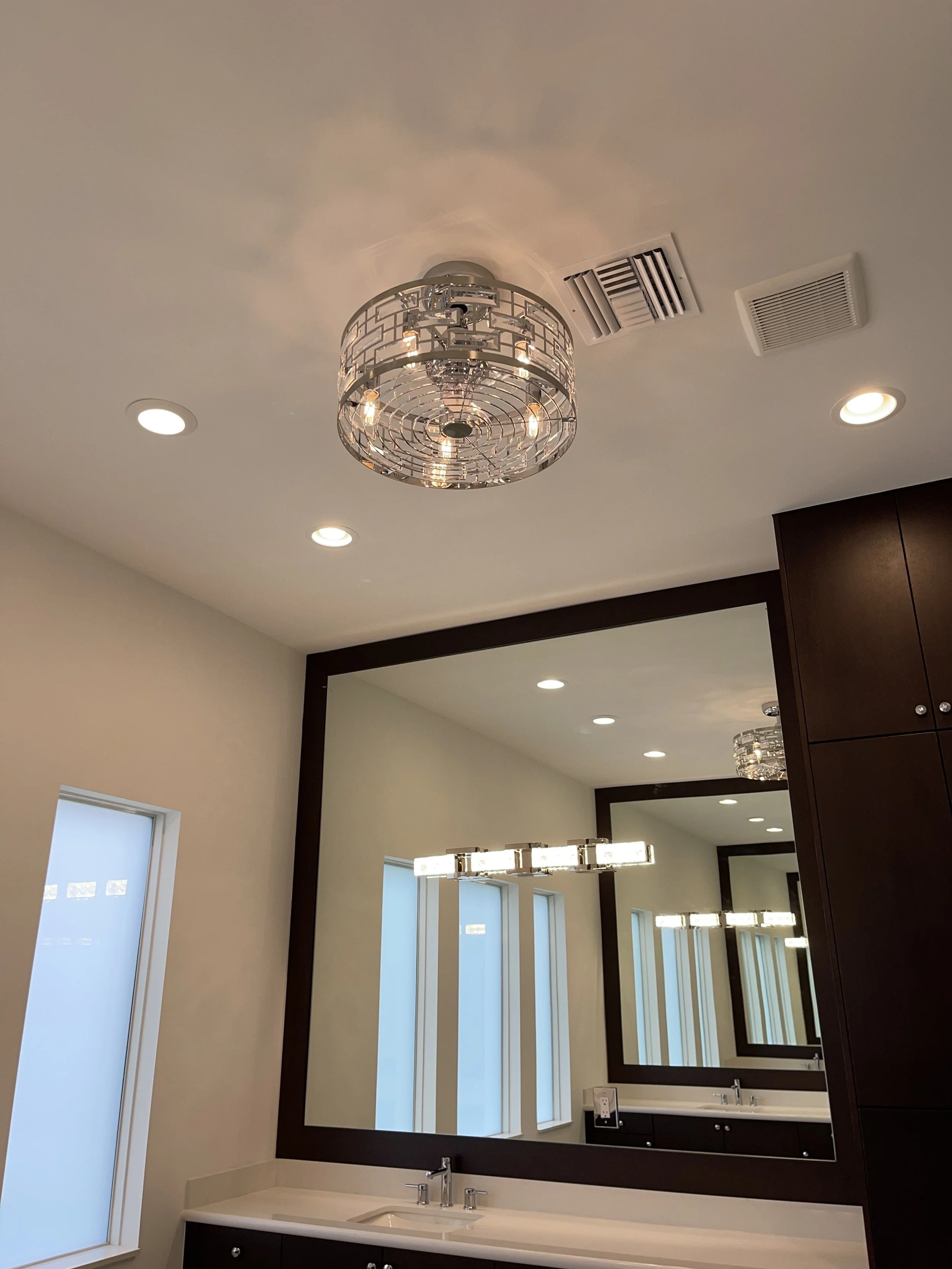Interior view of a modern bathroom with a large mirror, dark wood cabinetry, a white countertop with a sink, frosted windows, and a decorative ceiling light fixture.