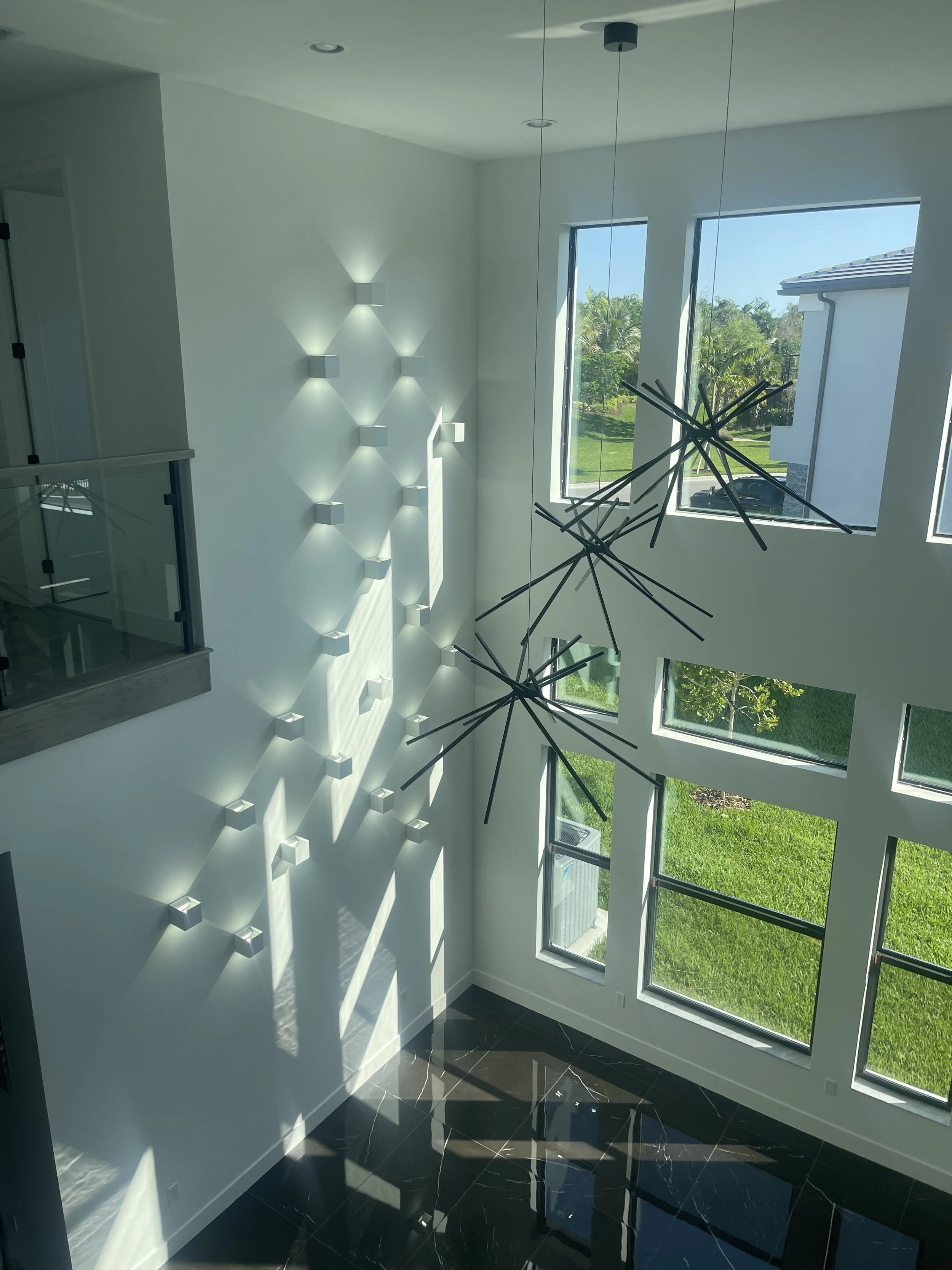 Modern living room with large windows, black chandelier with multiple rods, and wall-mounted light fixtures creating geometric shadows.