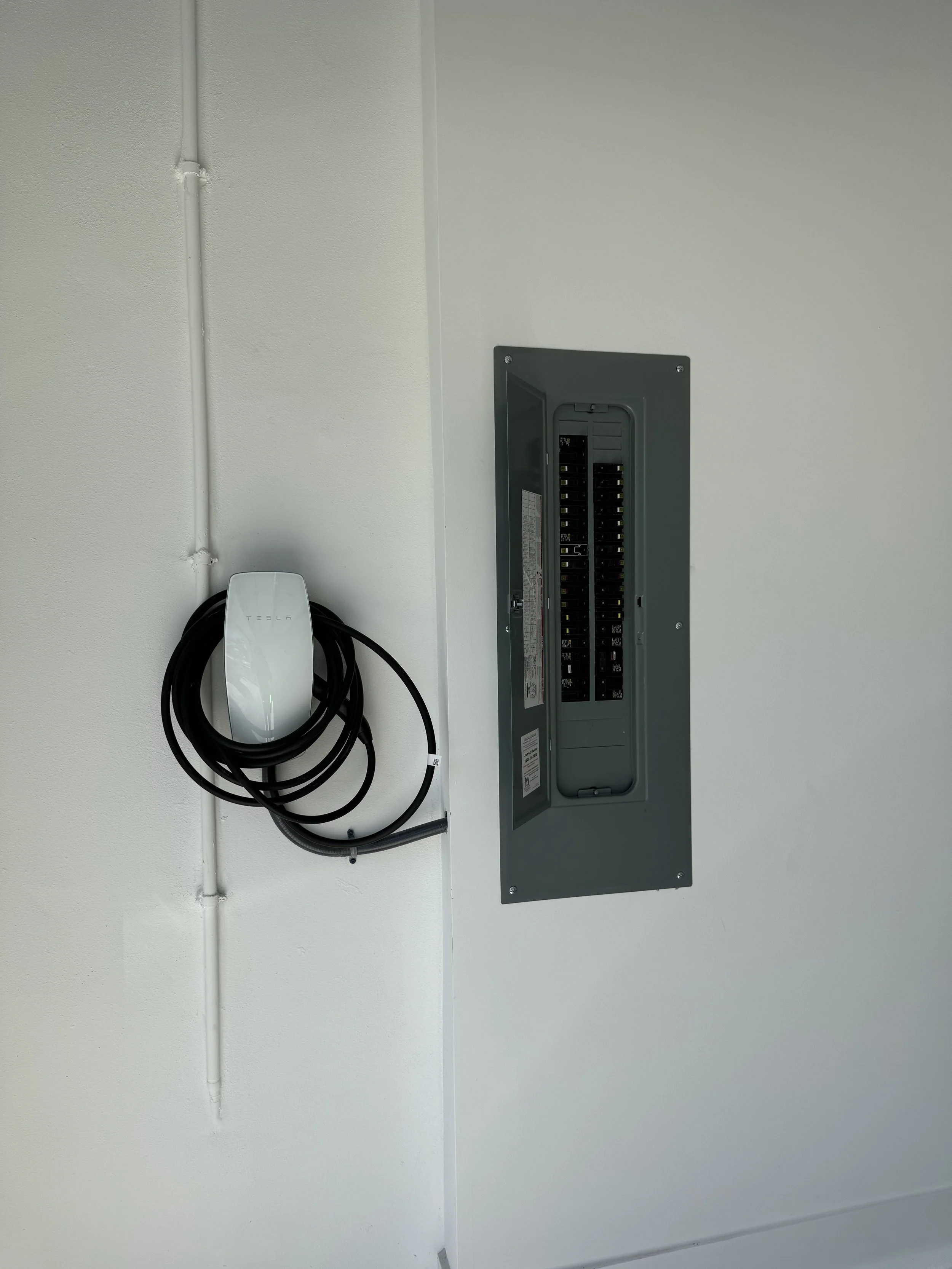 Electric vehicle charging station with a Tesla charging plug attached to a wall and an open electrical panel on a white wall.