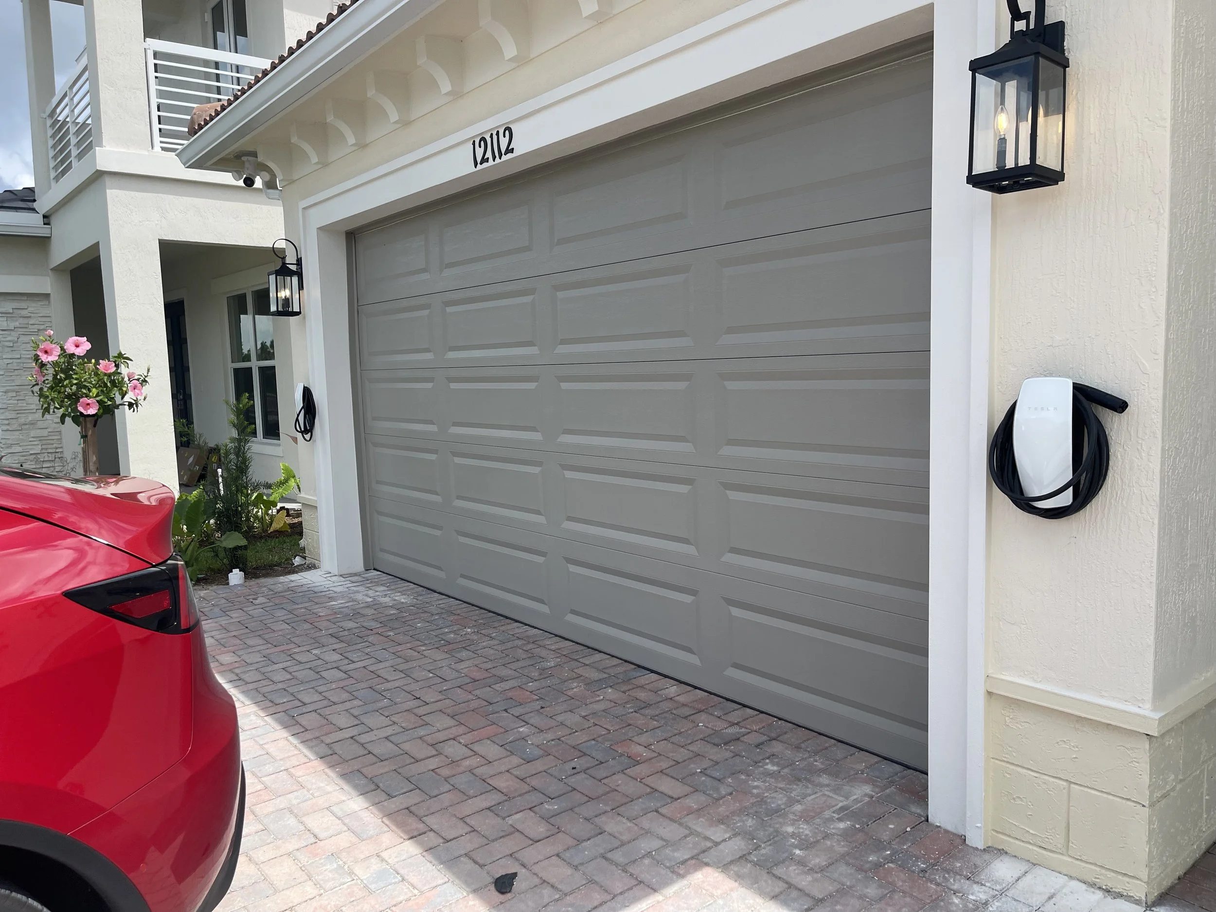 Close-up of a residential garage with a grey sectional garage door, a Tesla wall charger mounted on the cream-colored wall, and black exterior lantern-style lights. Part of a red car is visible in the driveway.