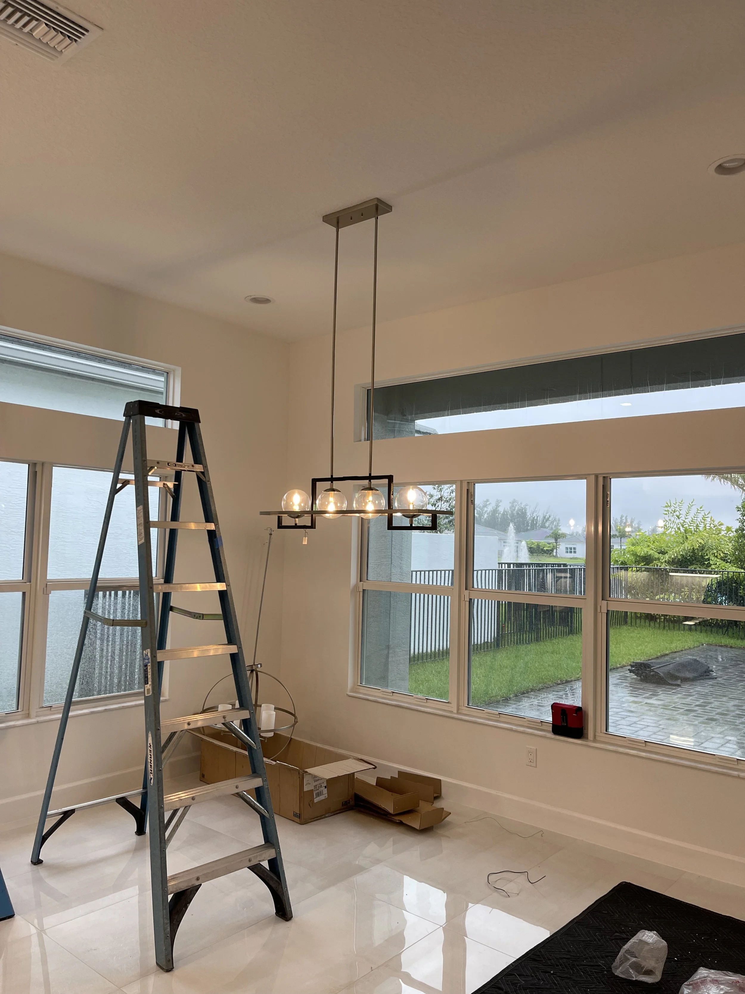 Interior of a room with large windows, a hanging light fixture with five bulbs, a ladder, and packing boxes on the floor.
