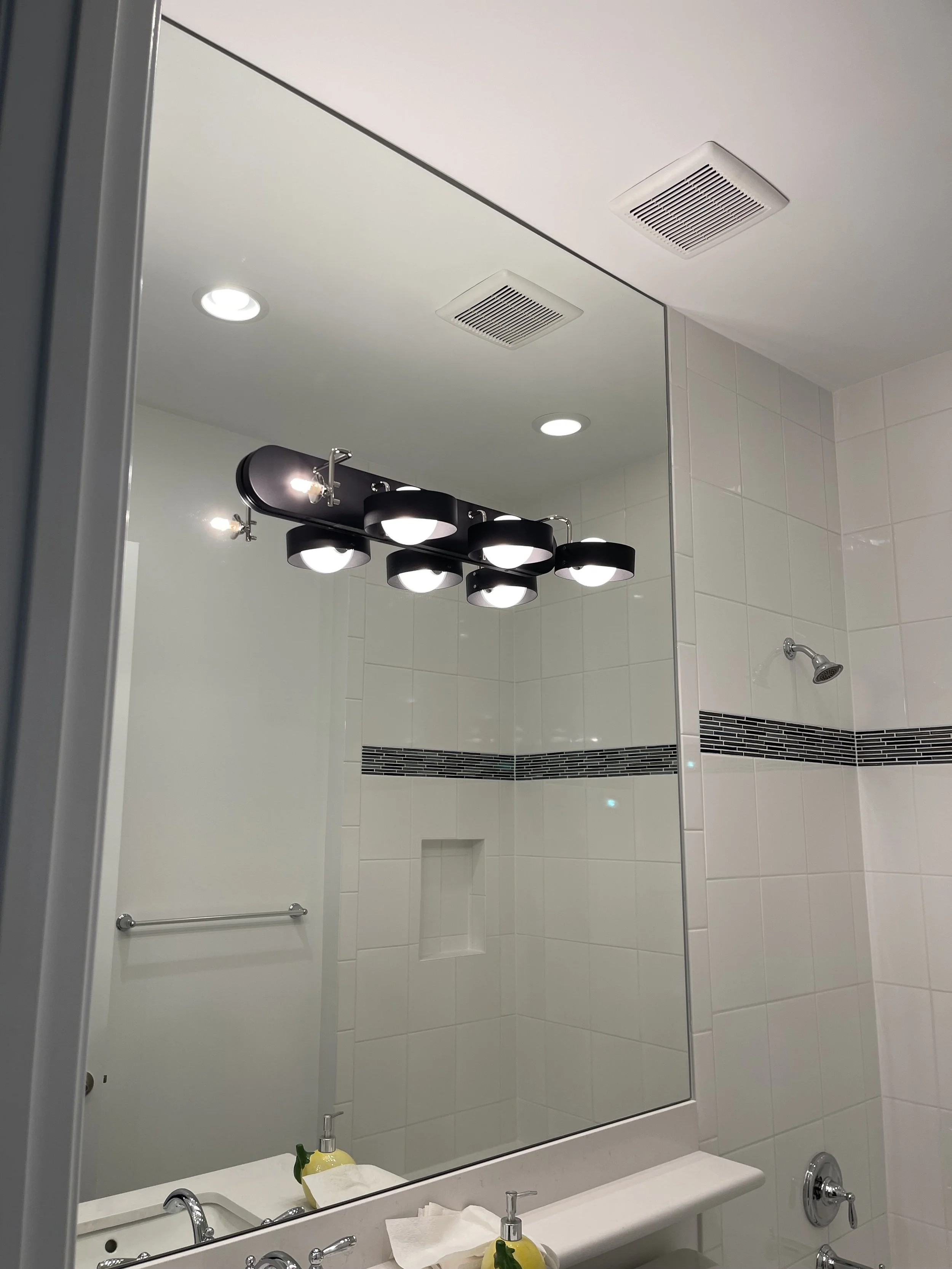 Bathroom mirror reflecting a vanity light fixture with five bulb lights, white tiled wall with black accent strip, showerhead, and bathroom sink with soap and paper towels.