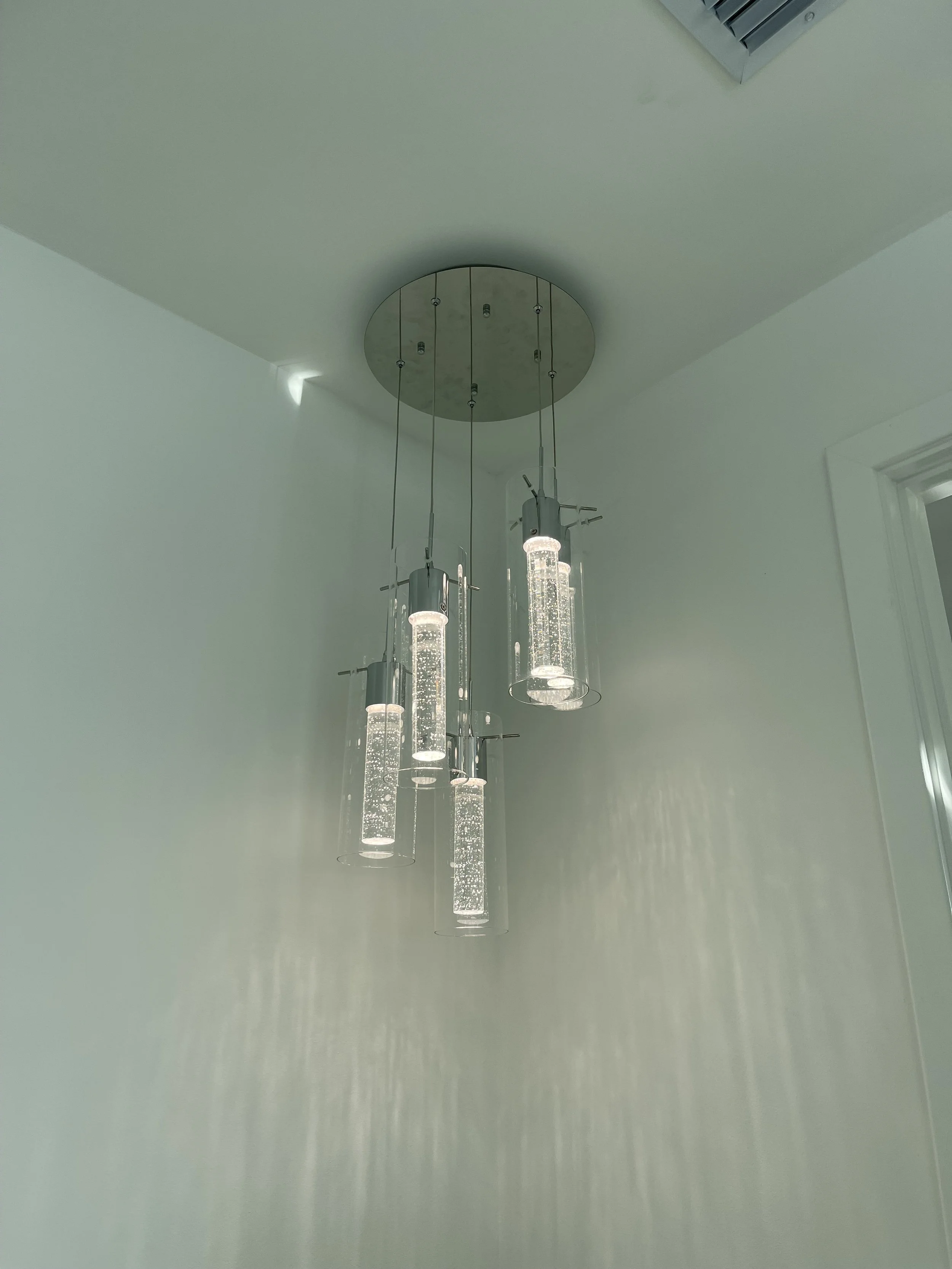 Modern hanging chandelier with multiple cylindrical glass tubes filled with bubbles, mounted on a round ceiling fixture, in a room with white walls and ceiling.