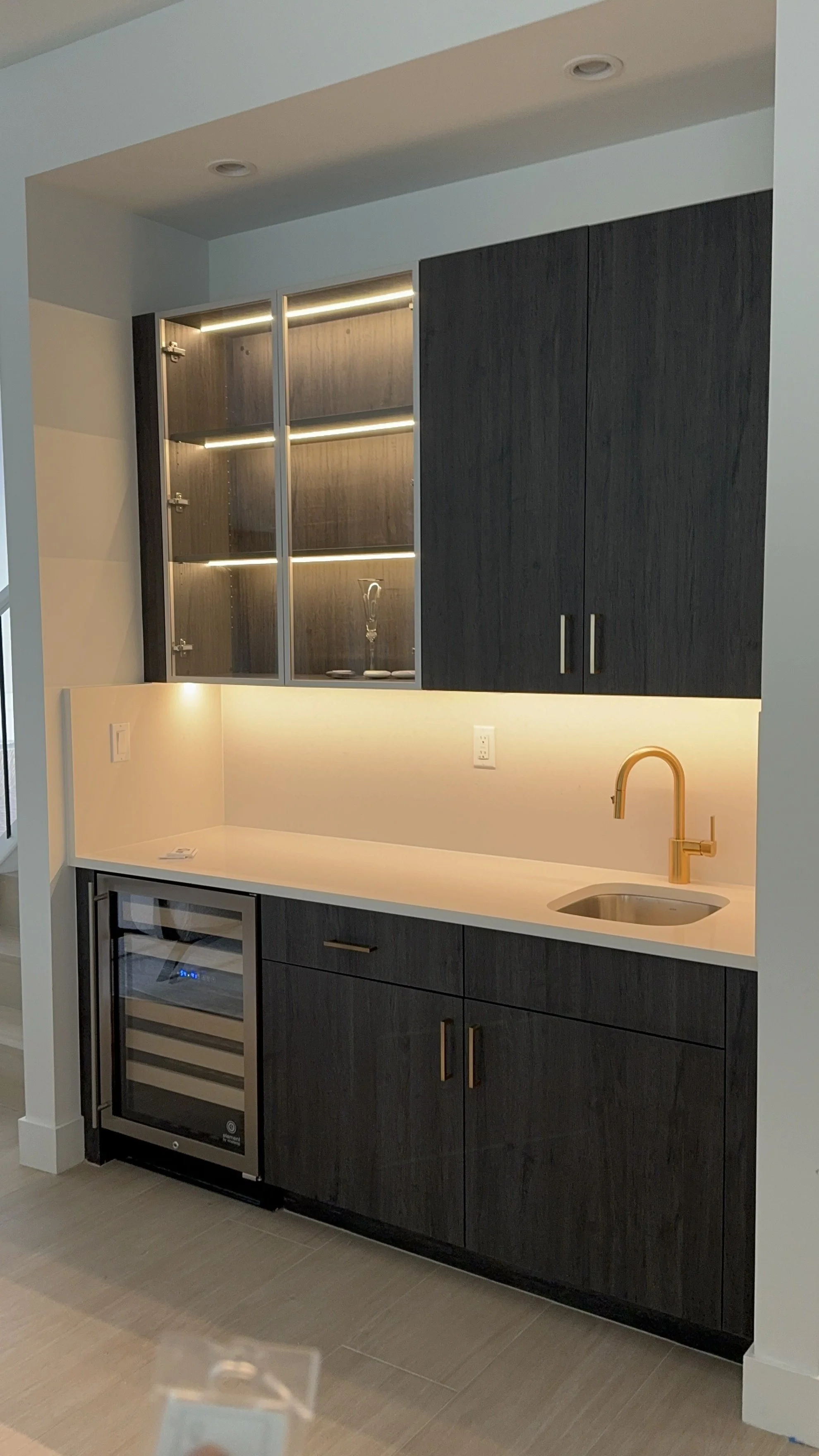 Modern wet bar with black wooden cabinets, a mini fridge, a white countertop, a gold faucet, and glass display shelves with LED lighting.