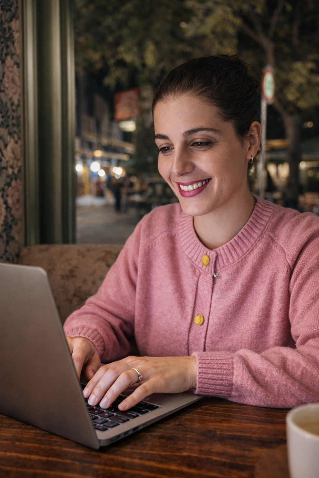 A woman with brown hair tied back, wearing a pink sweater with yellow buttons, smiling while using a laptop at a cafe in the evening, with outdoor seating and trees visible through the window.