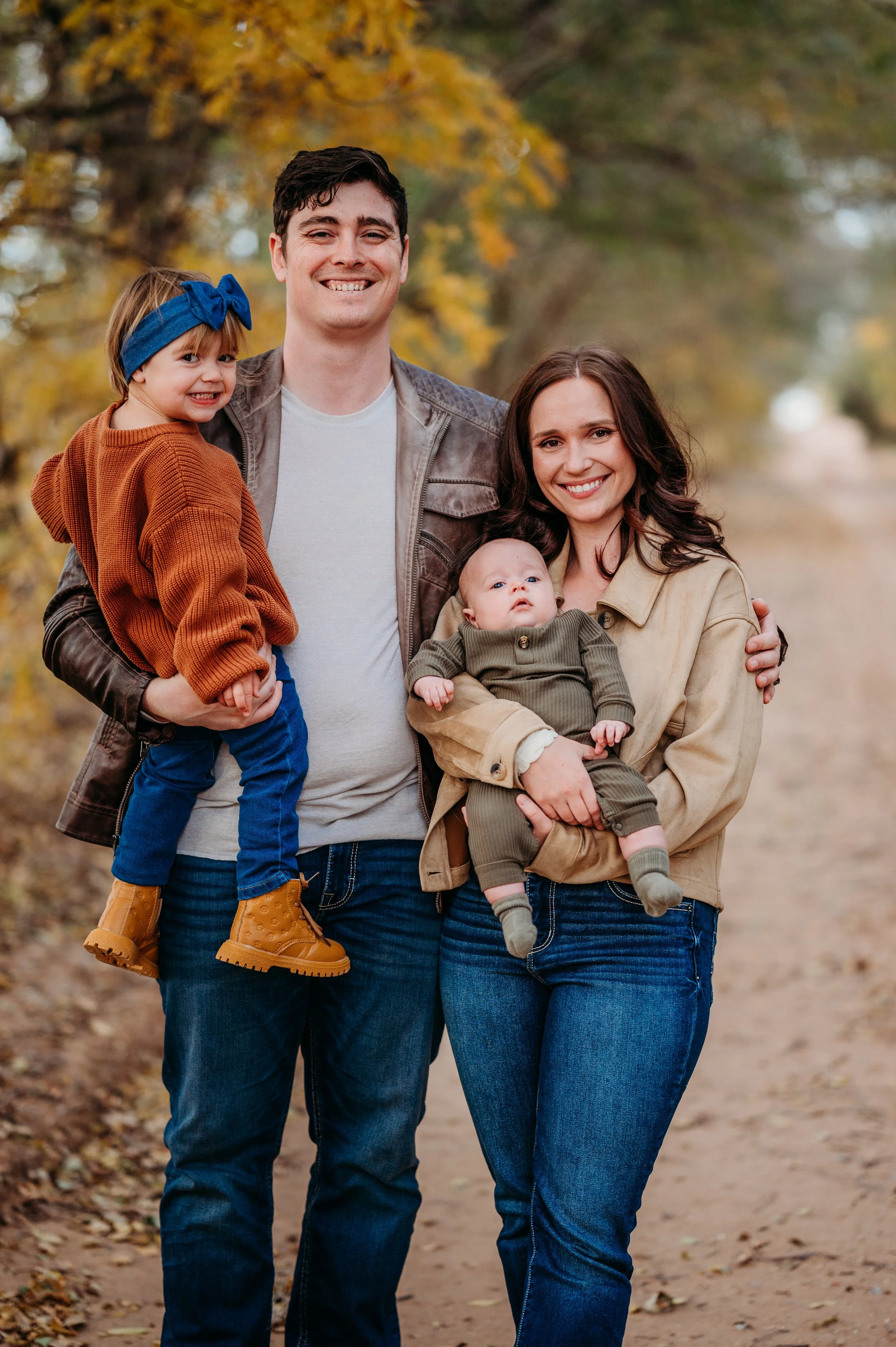 A family of four standing outdoors on a fall day. The father is holding a young girl, and the mother is holding a baby. They are all smiling and dressed in casual clothing, with autumn leaves in the background.