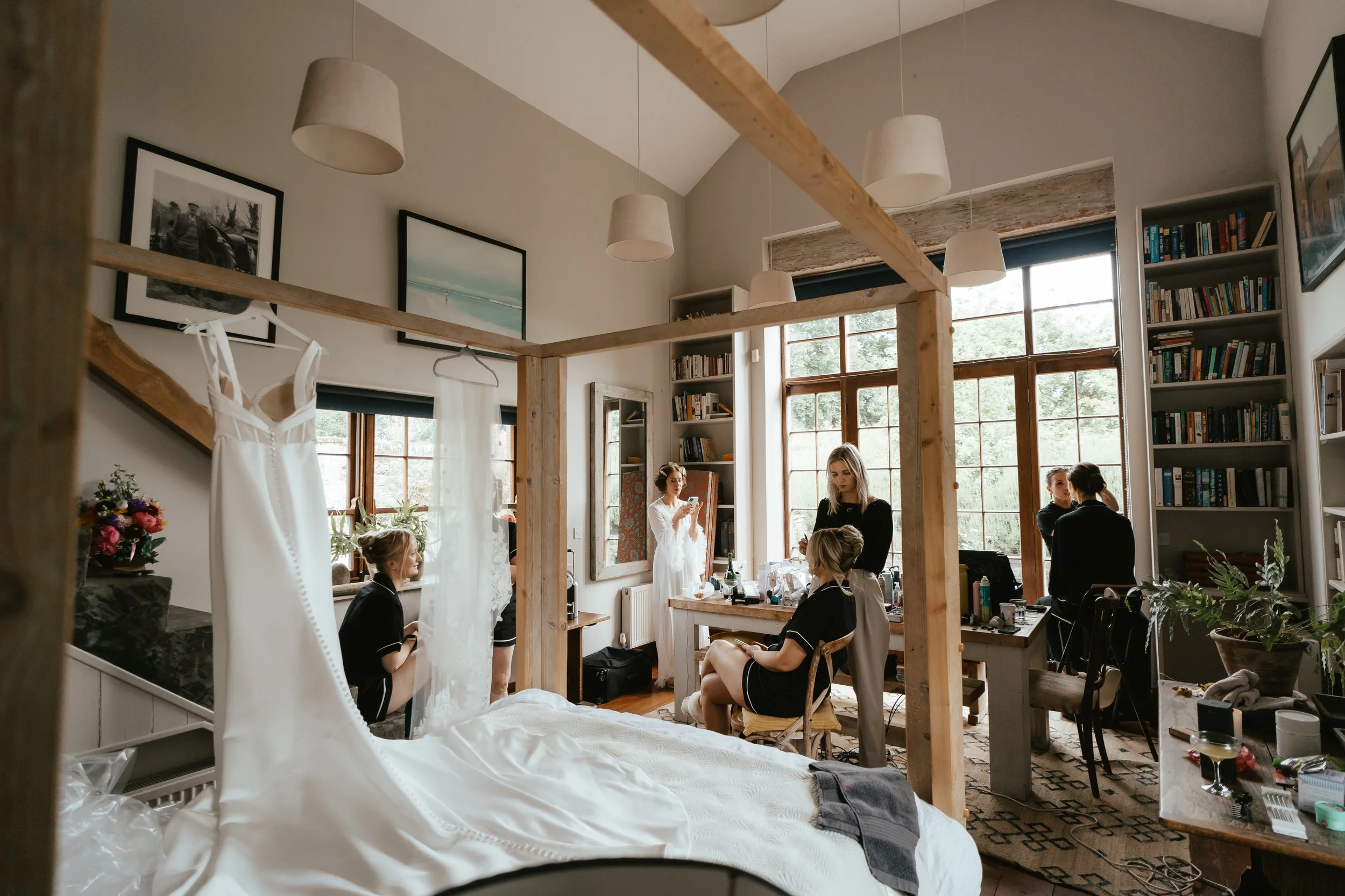 Women preparing for a wedding in a sunlit room with large windows, bookshelves, and a wedding dress hanging on a bed.