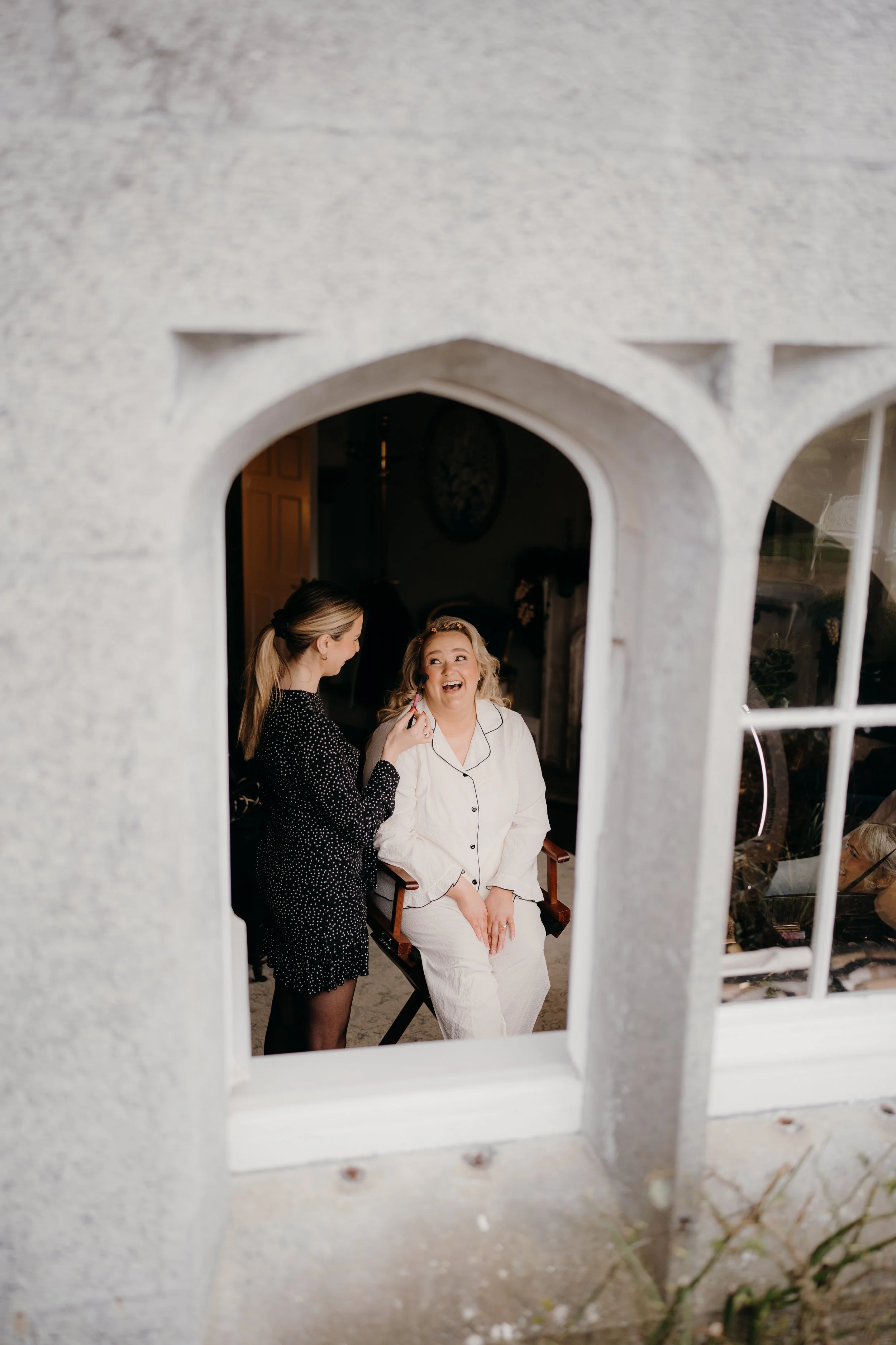 A woman sitting in a chair having her makeup done by a makeup artist, seen through a decorative window opening.