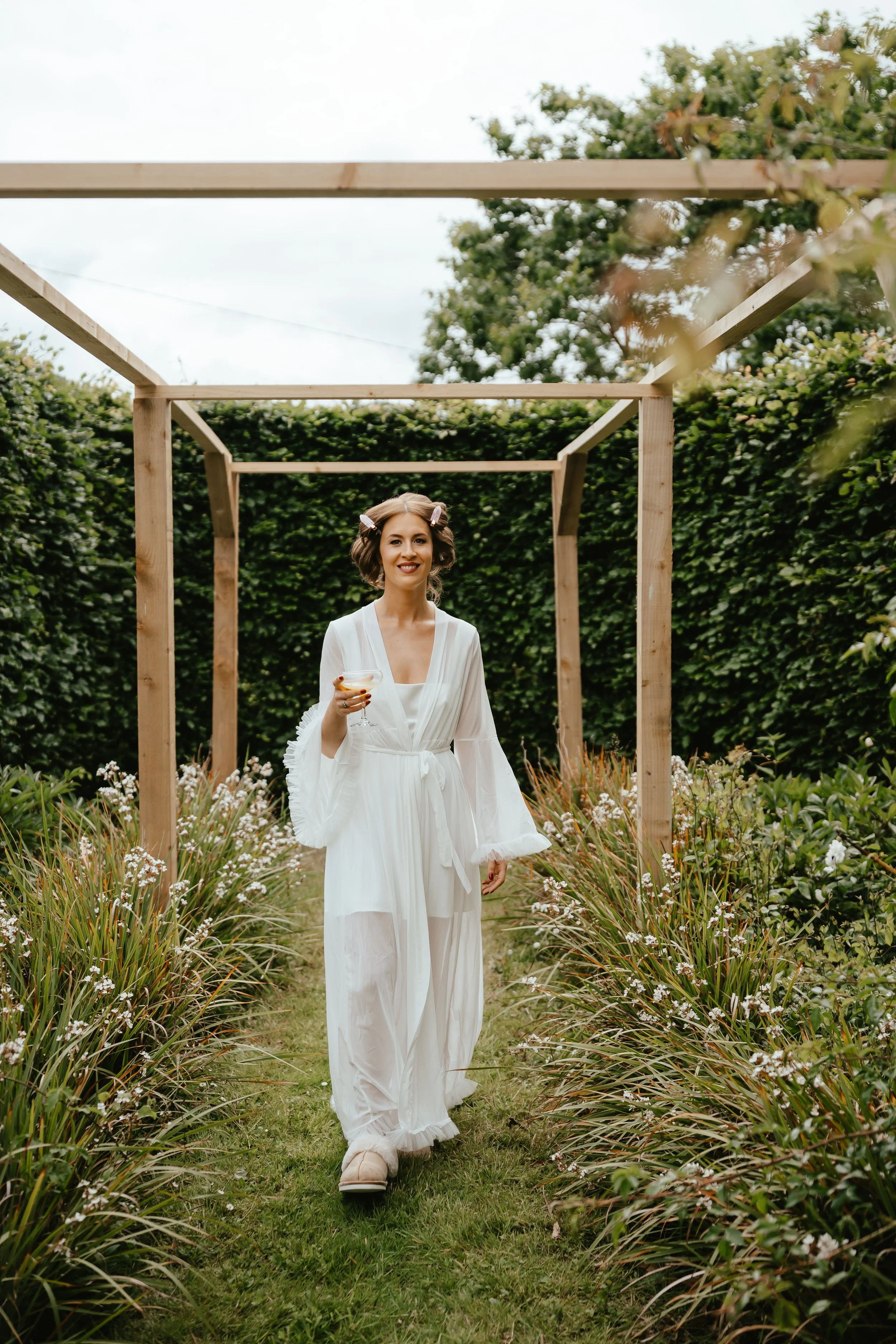 A woman in a white robe and slippers walking through a garden path holding a drink, with a wooden frame structure overhead and green hedges and plants on either side.