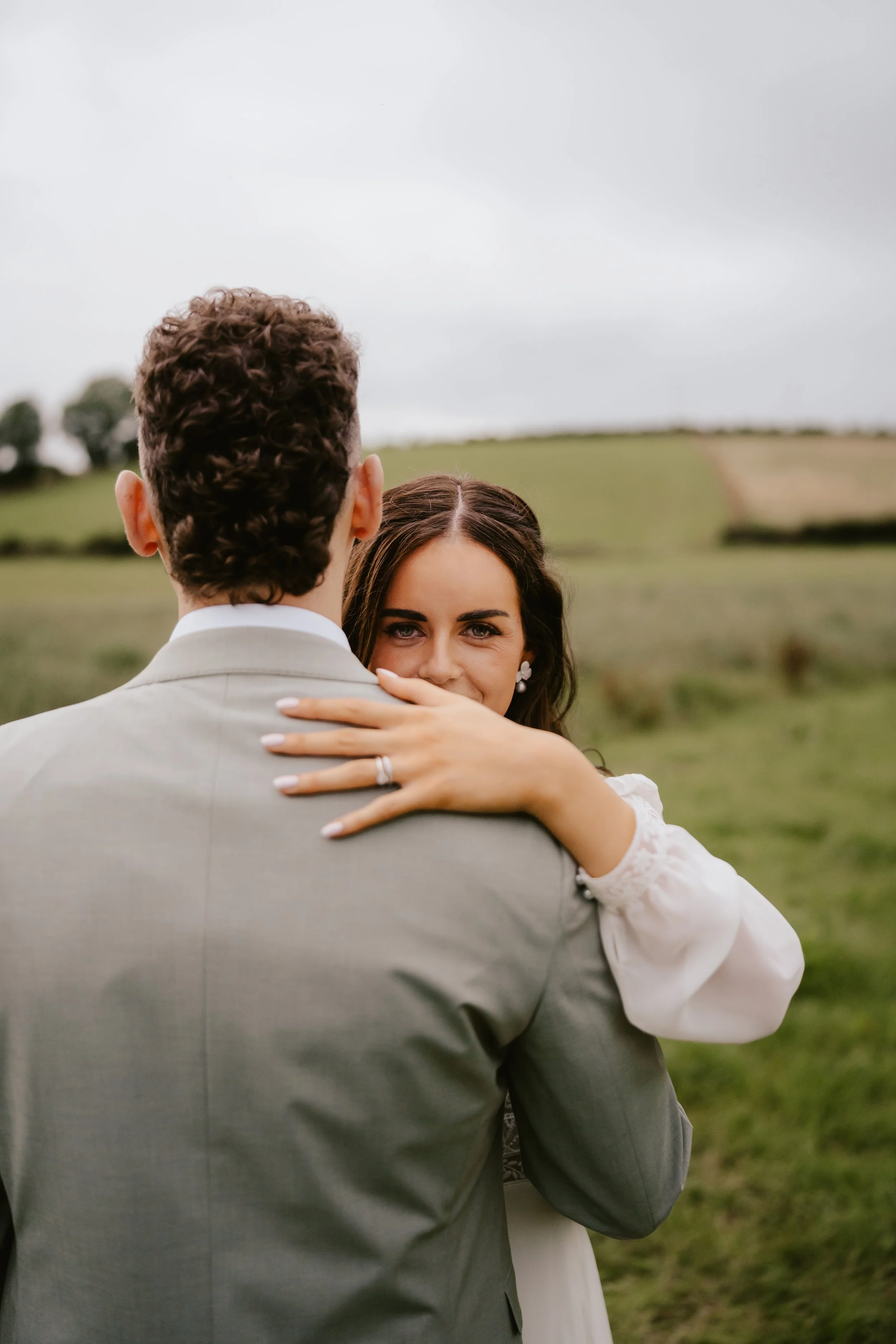 A woman hugging a man outdoors in a grassy field under a cloudy sky, with the woman's face visible as she gazes at the camera.