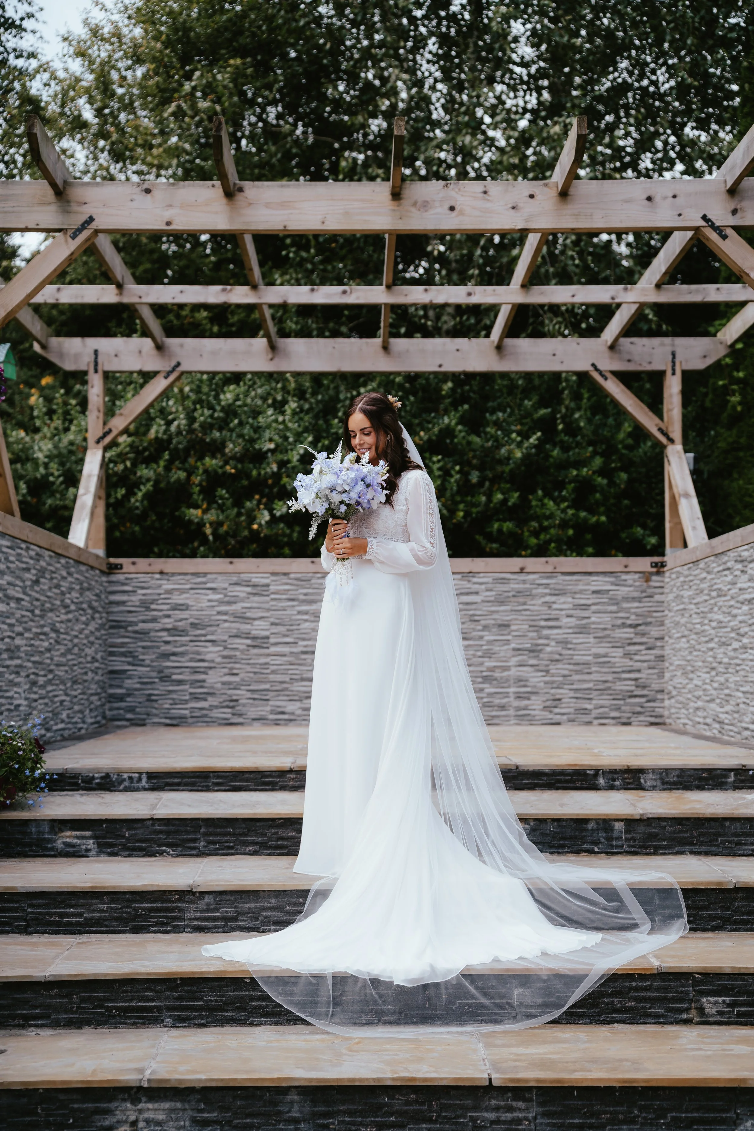 A bride in a white wedding gown holding a bouquet of white and purple flowers, standing on a wooden stage outdoors with a pergola structure and green trees in the background.