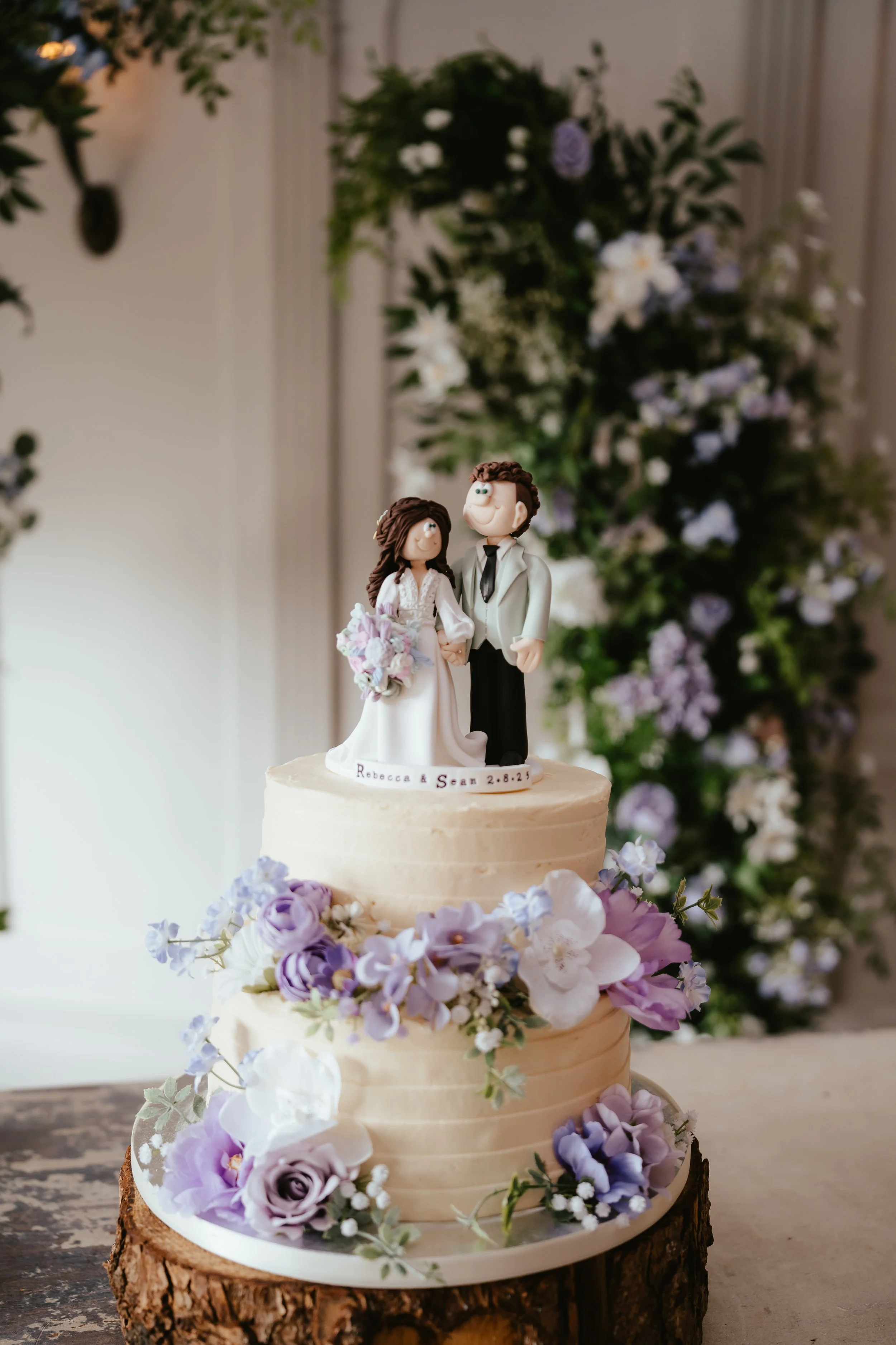 A wedding cake with purple and white flowers and a topper of a cartoon bride and groom.