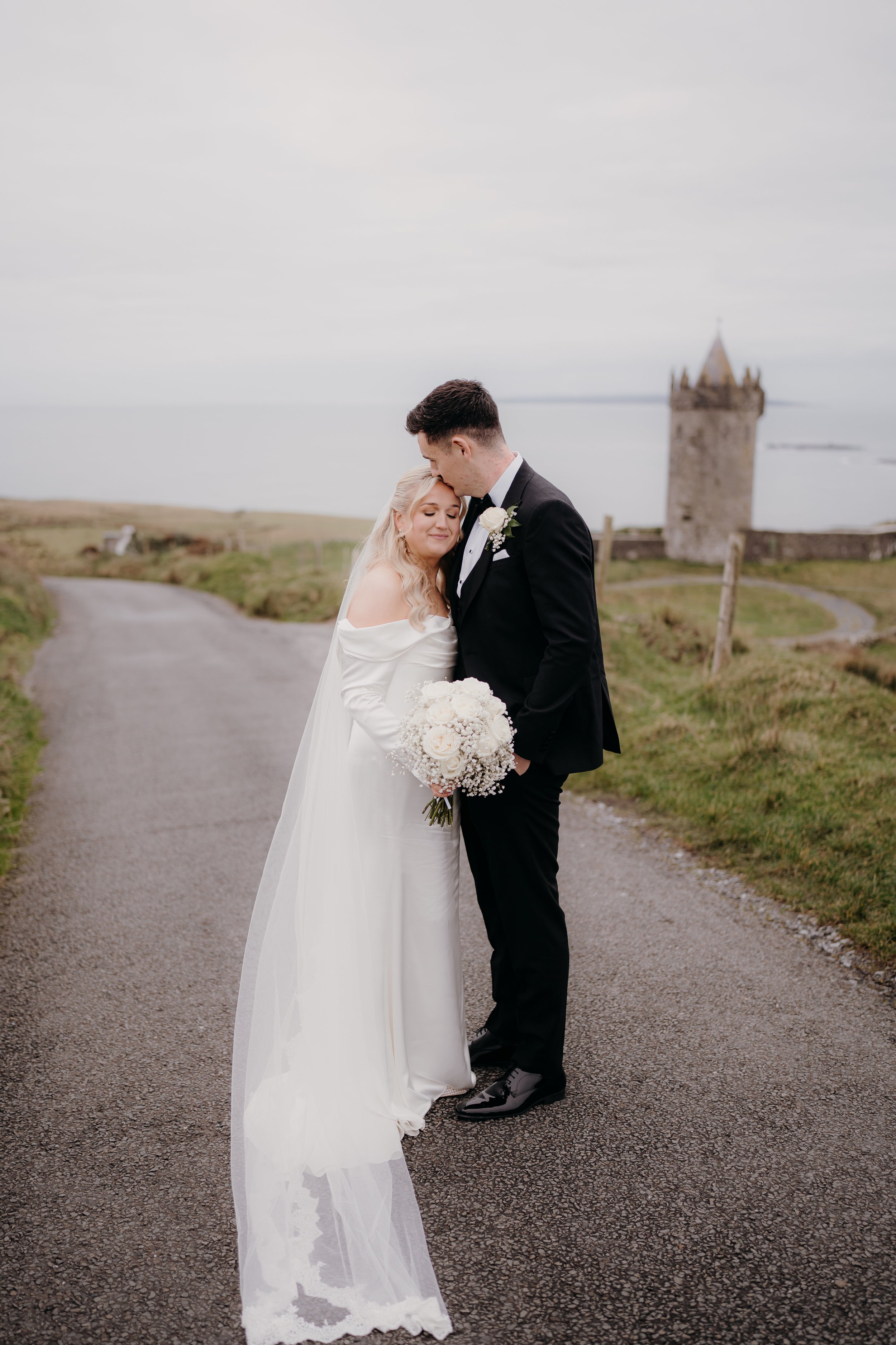 A bride and groom standing close together on a rural road, with a castle in the background, during their wedding day.