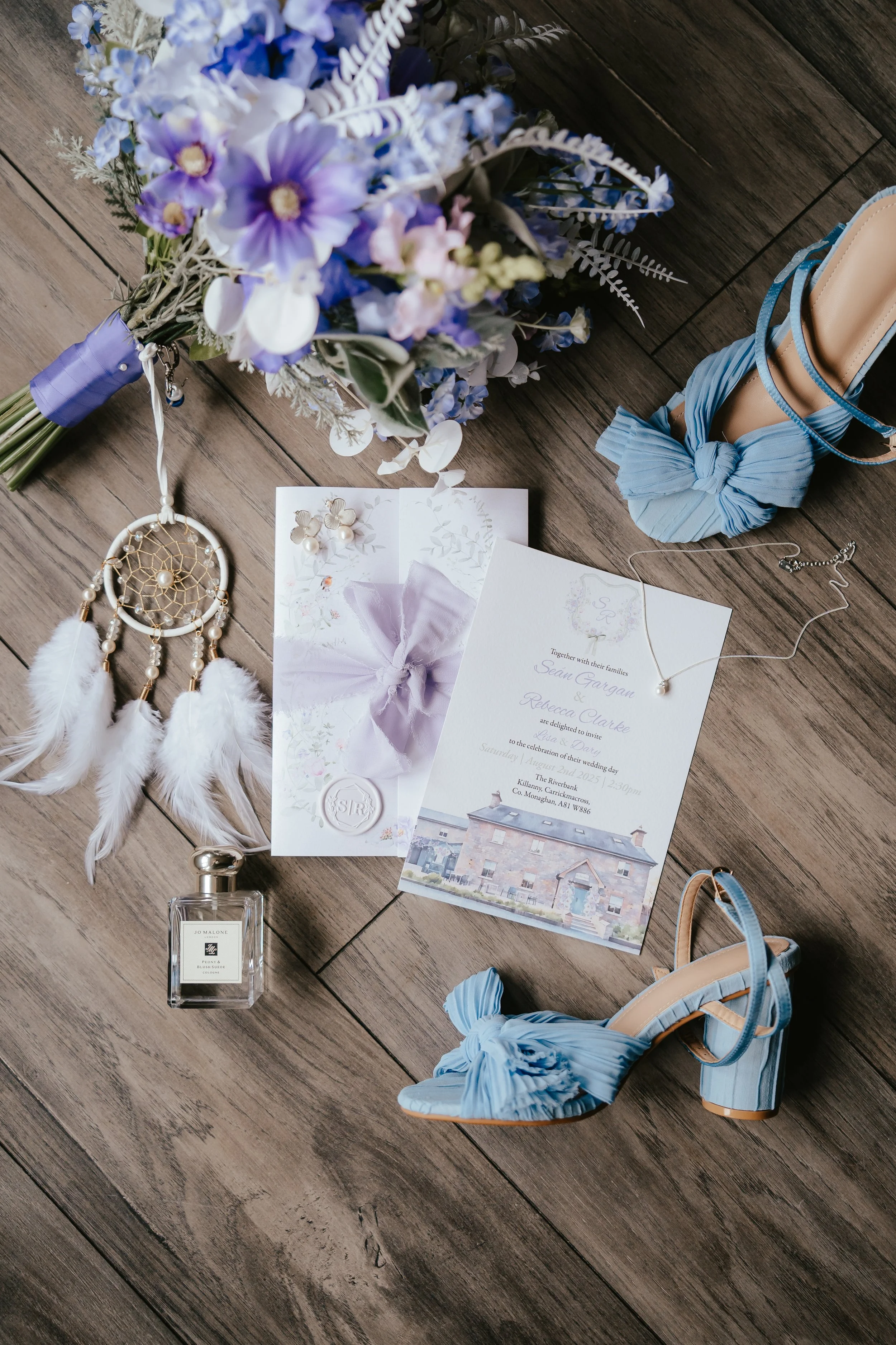Wedding invitation, bouquet of purple and white flowers, blue high-heeled shoes with bow, white dreamcatcher with feathers, perfume bottle, necklace with pearl, and a ring tied with purple ribbon on a wooden floor.