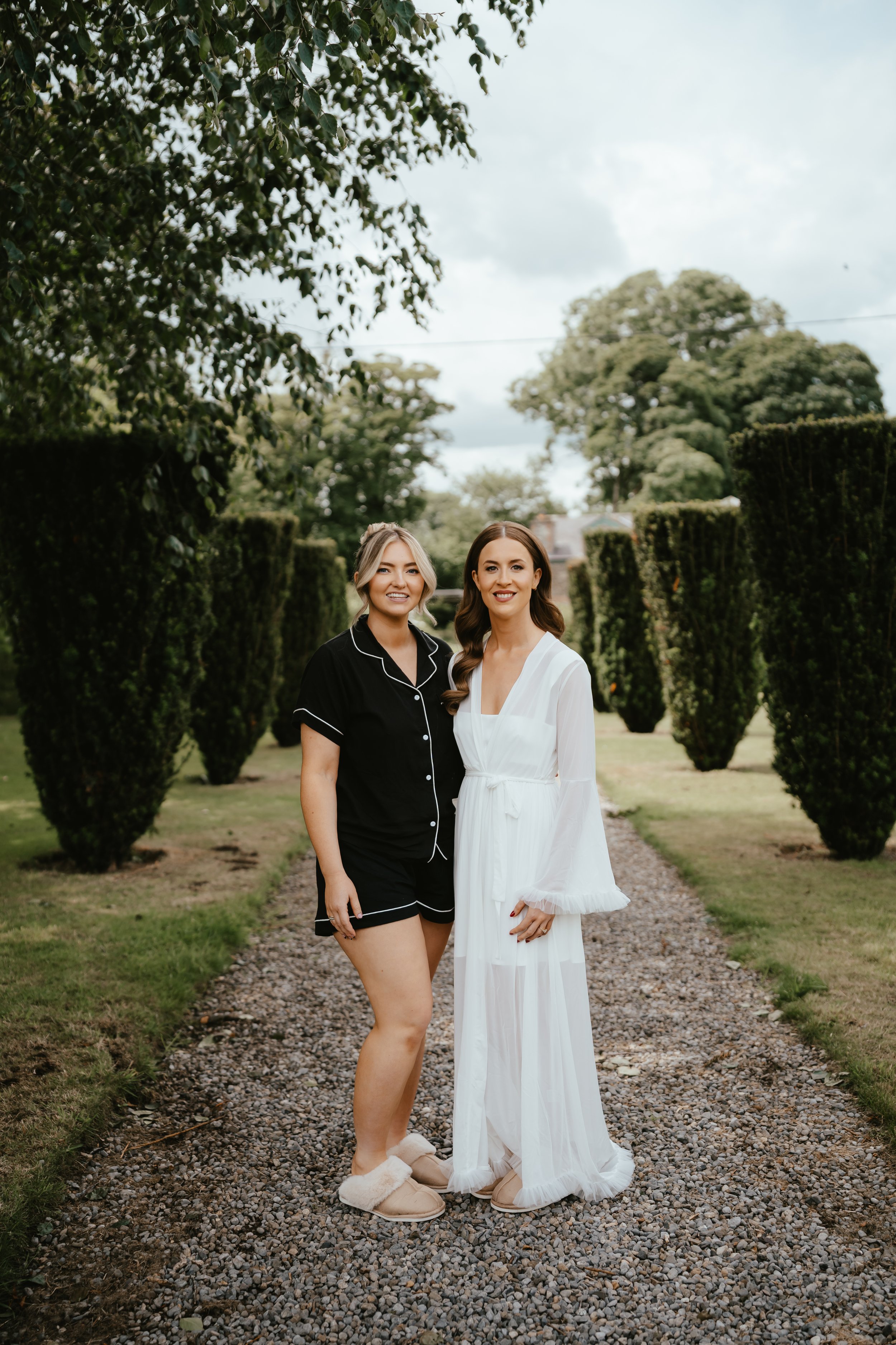 Two women standing on a gravel path, smiling at the camera, surrounded by tall, lush green trees and bushes, with a cloudy sky overhead.