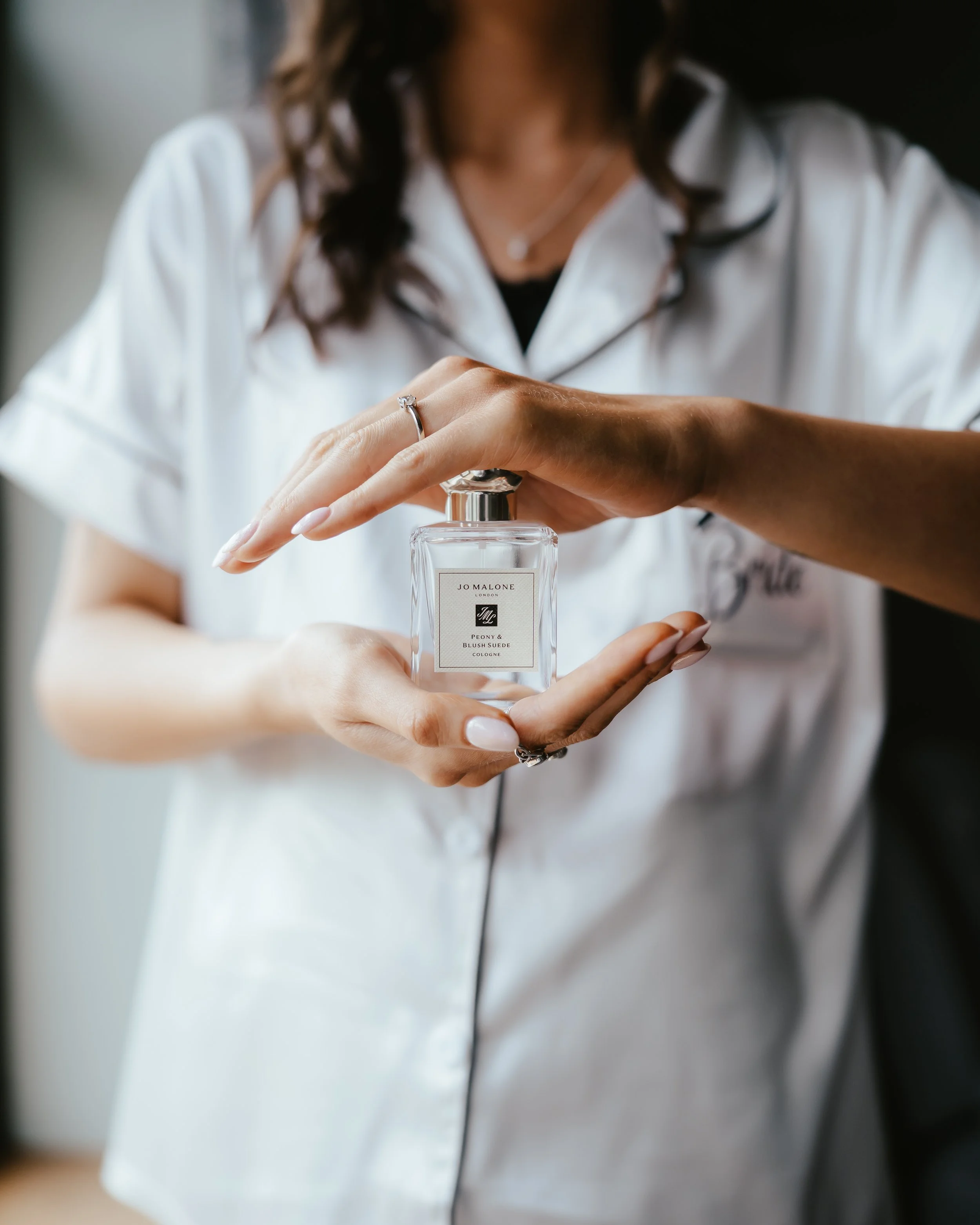 Person holding a perfume bottle labeled 'Jo Malone London Peony & Blush Suede' with both hands, wearing a white shirt.