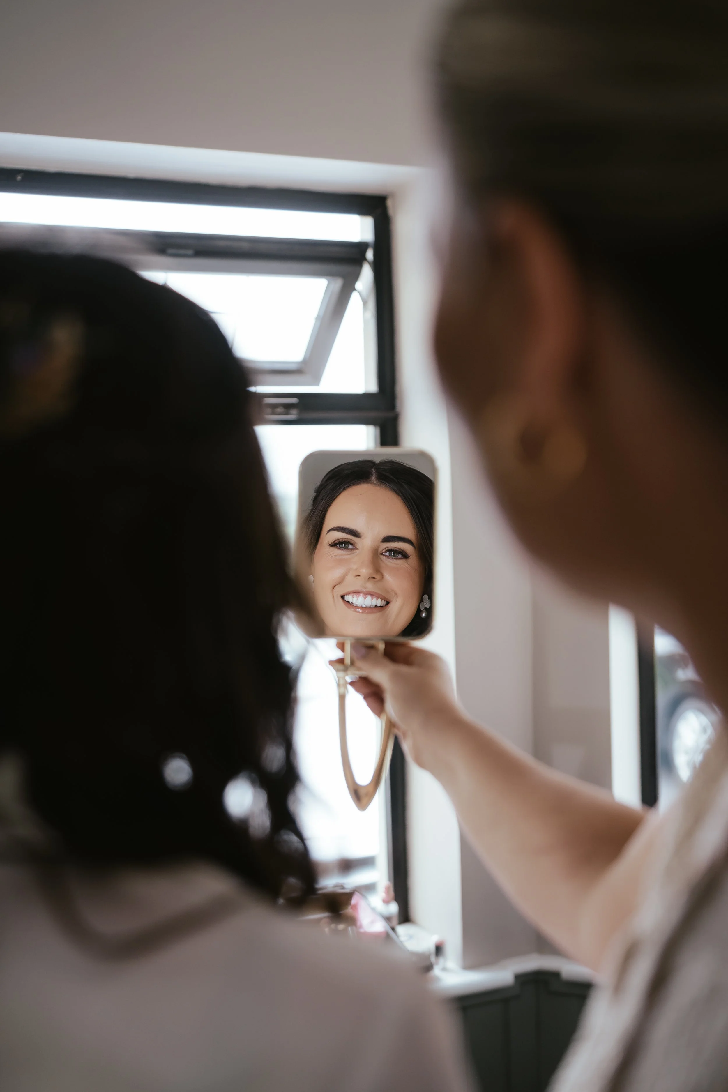 Two women are looking at a small mirror; the woman in the mirror is smiling, while the other woman holds a brush.