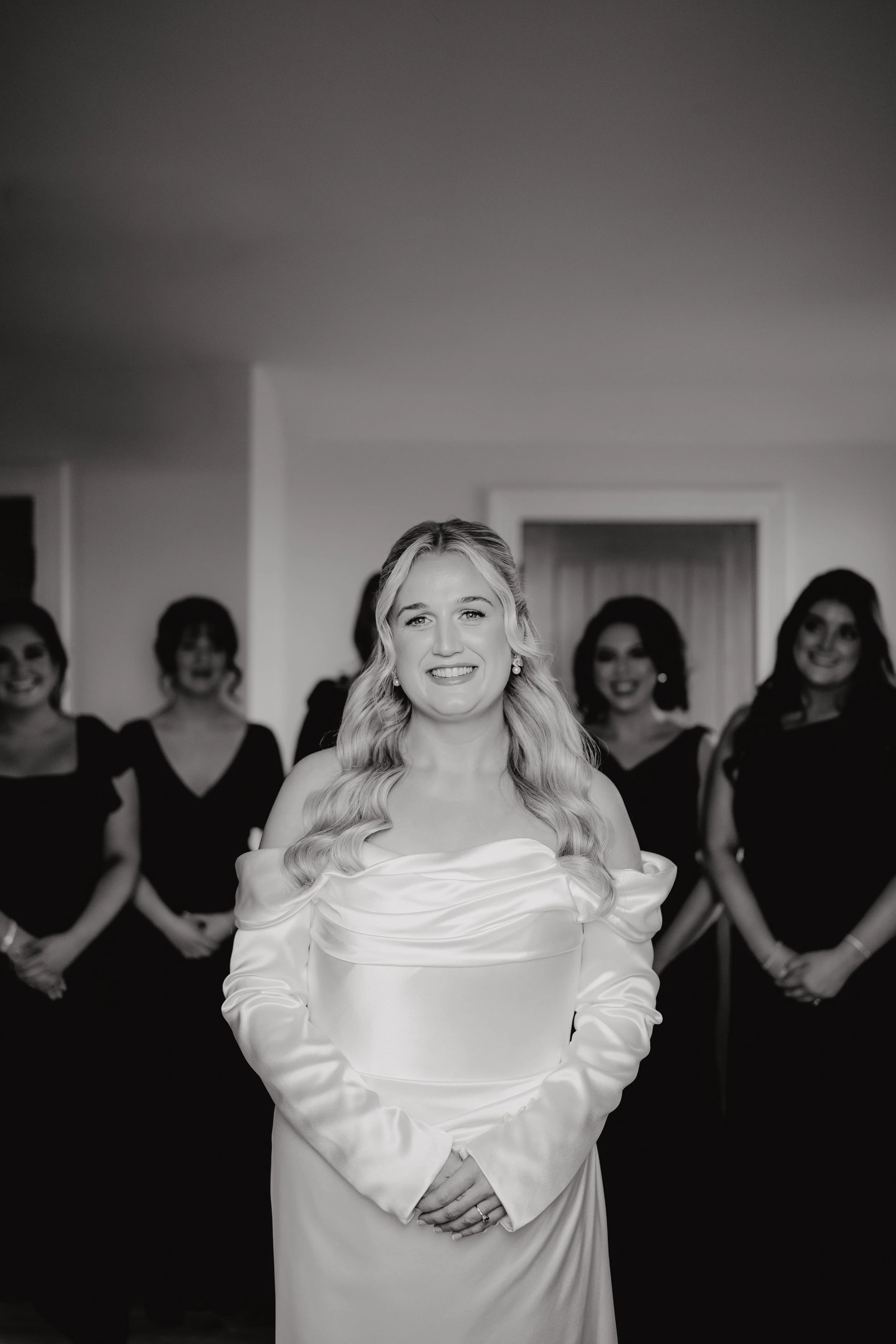 A smiling woman in a wedding dress stands in the foreground with four women in black dresses behind her in a room.