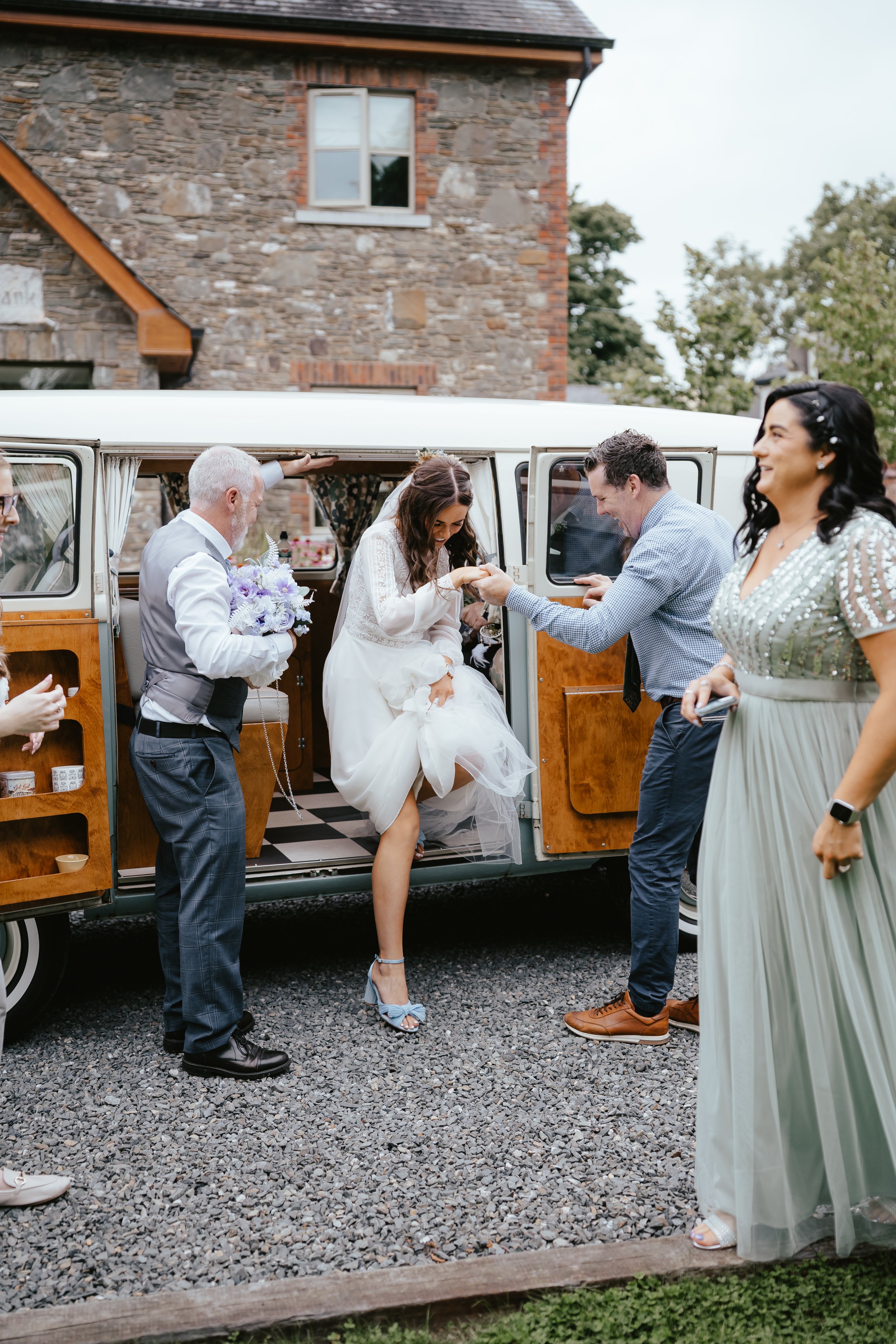 A bride in a white dress and high heels stepping out of a vintage van, smiling and being handed a flower by a man, with several people around her celebrating.