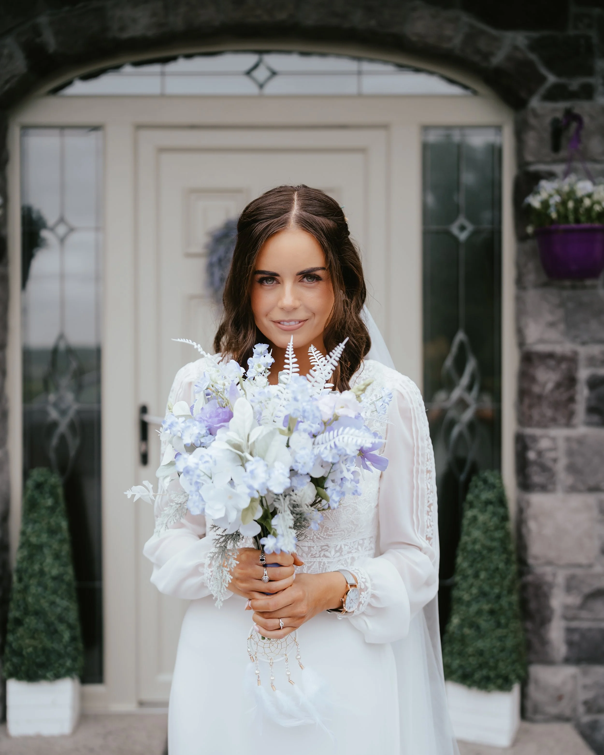 Bride holding a bouquet of white and light purple flowers standing in front of a door.