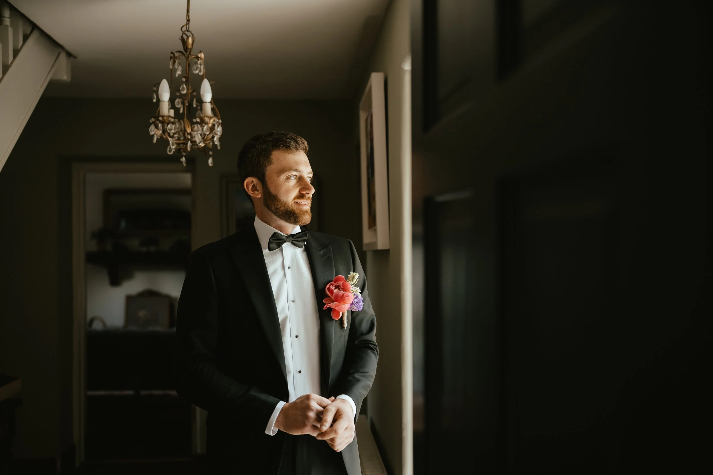 A man in a black tuxedo with a bow tie and boutonnière stands by a window looking outside in a dimly lit room, with a chandelier hanging from the ceiling.