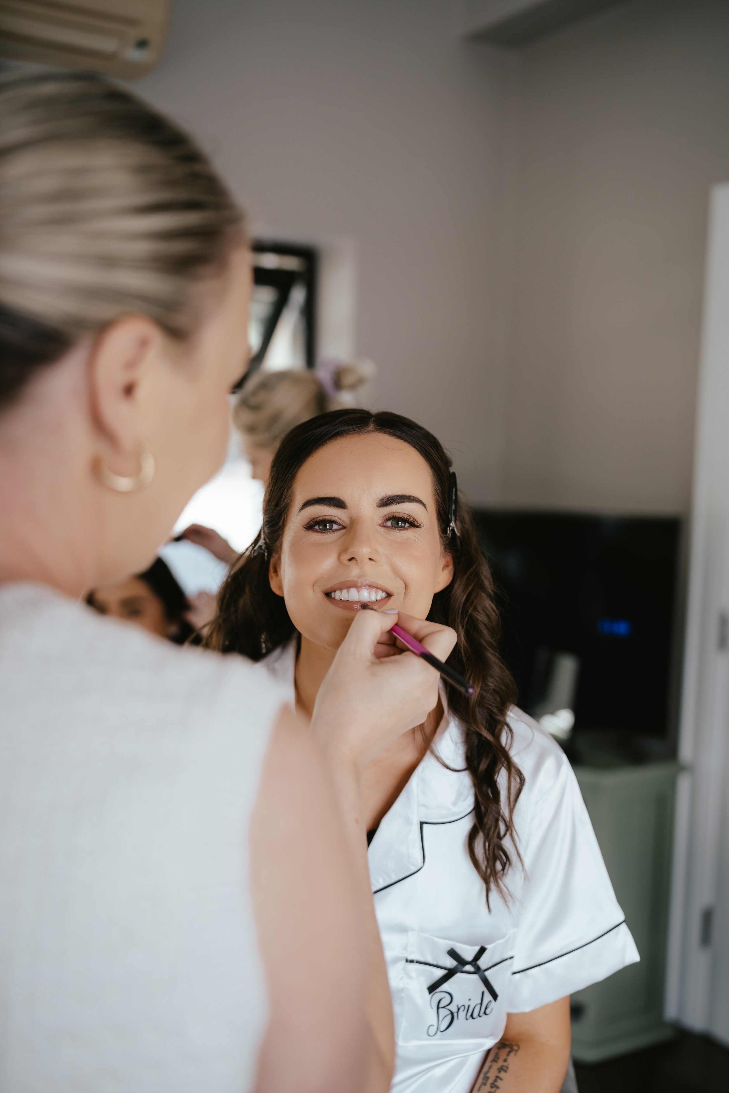 A bride with long dark hair is smiling as a makeup artist applies lipstick. The bride is wearing a satin pajama top with the word 'Bride' embroidered on it. In the background, another woman is partially visible.